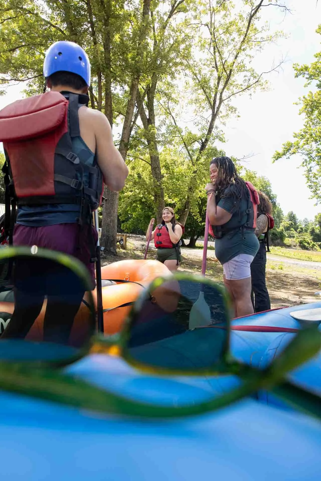 Group of four people preparing for kayaking outdoors, with the focus on three women and one man wearing life jackets and holding paddles, near parked kayaks under a tree-lined sky.