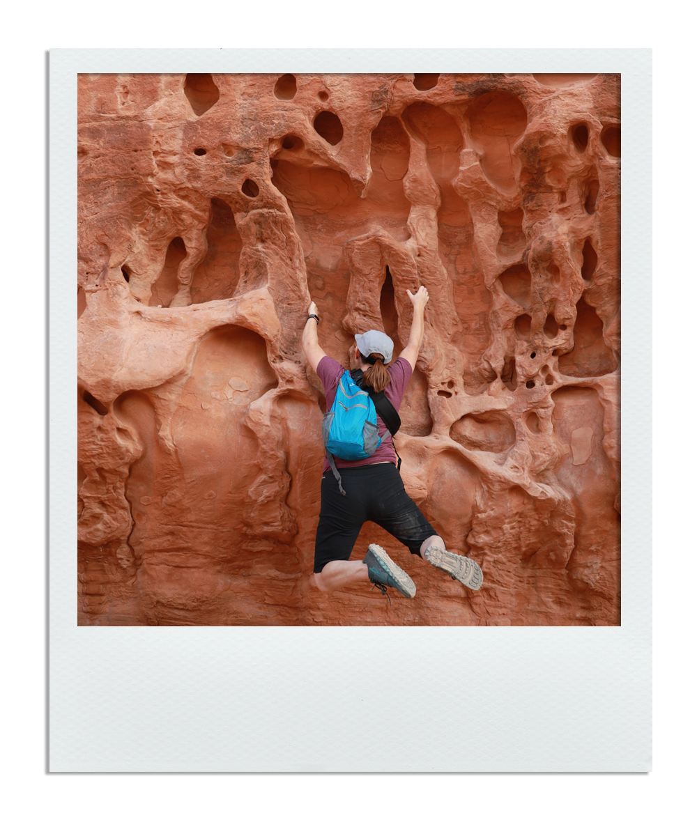 Person rock climbing on a textured red sandstone wall with multiple holes, wearing athletic clothing and a backpack.