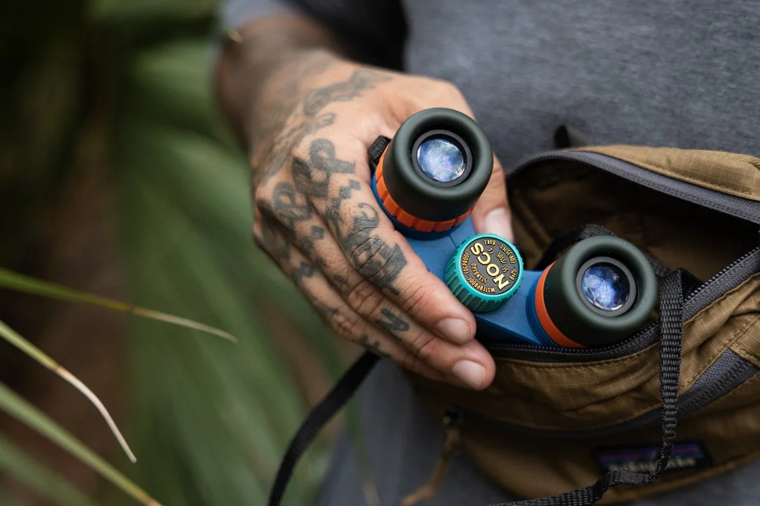 Person holding a compact blue and black binocular with orange accents in a brown backpack.