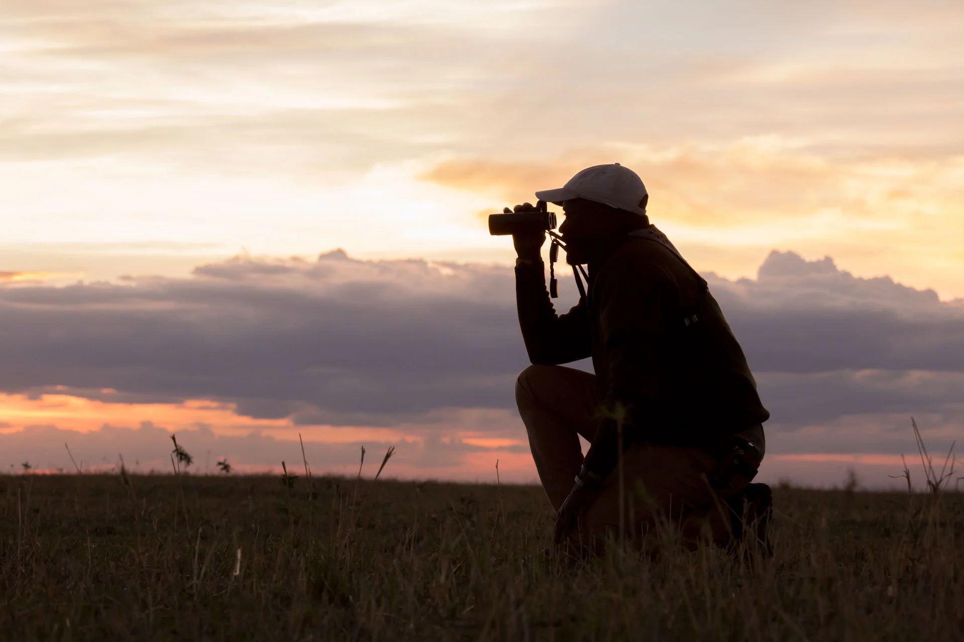 Silhouette of a person kneeling outdoors, looking through binoculars during sunset or sunrise with clouds and a colorful sky in the background.
