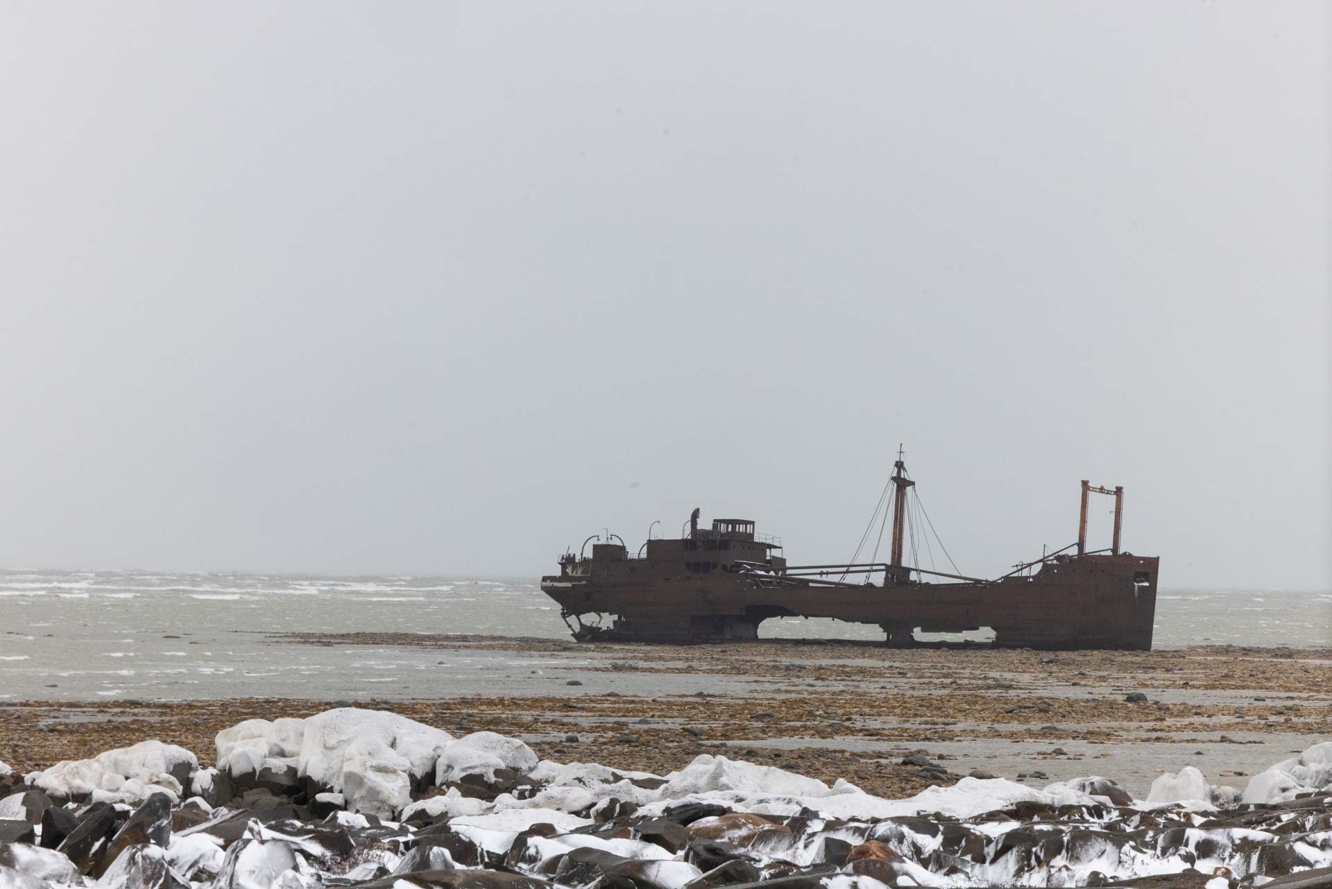 Old, derelict shipwreck on a snowy, rocky shoreline in a foggy, desolate environment.