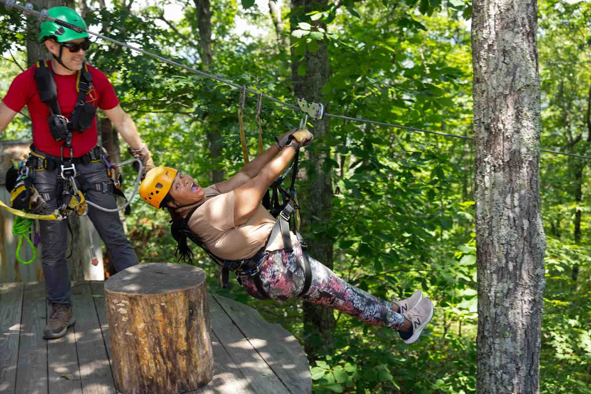 A woman in a yellow helmet and floral leggings is zip-lining through a forest, while a man in a green helmet and red shirt, equipped with safety harnesses, assists her from a wooden platform.