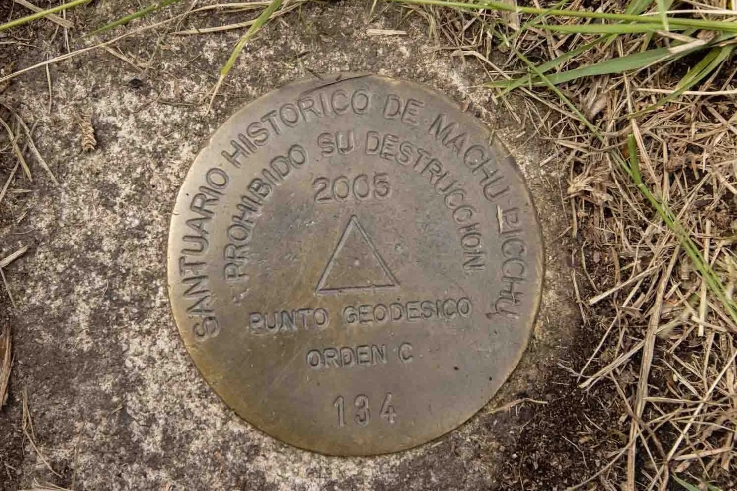 A geodesic survey marker embedded in ground surrounded by grass and dirt, with inscription in Spanish indicating its historical significance.