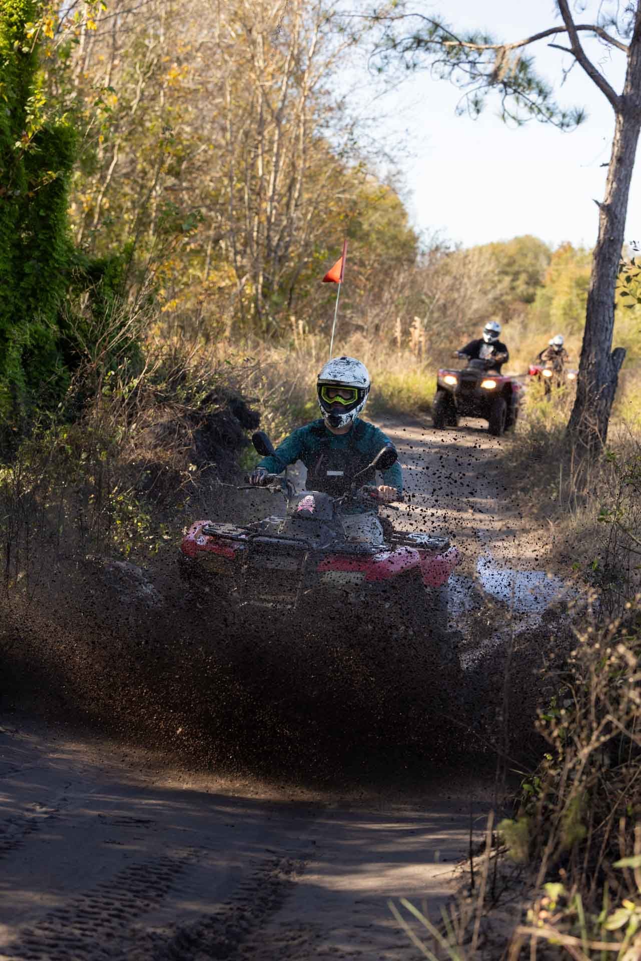 A person riding an ATV through a muddy trail in a wooded area, with two more riders on ATVs in the background.