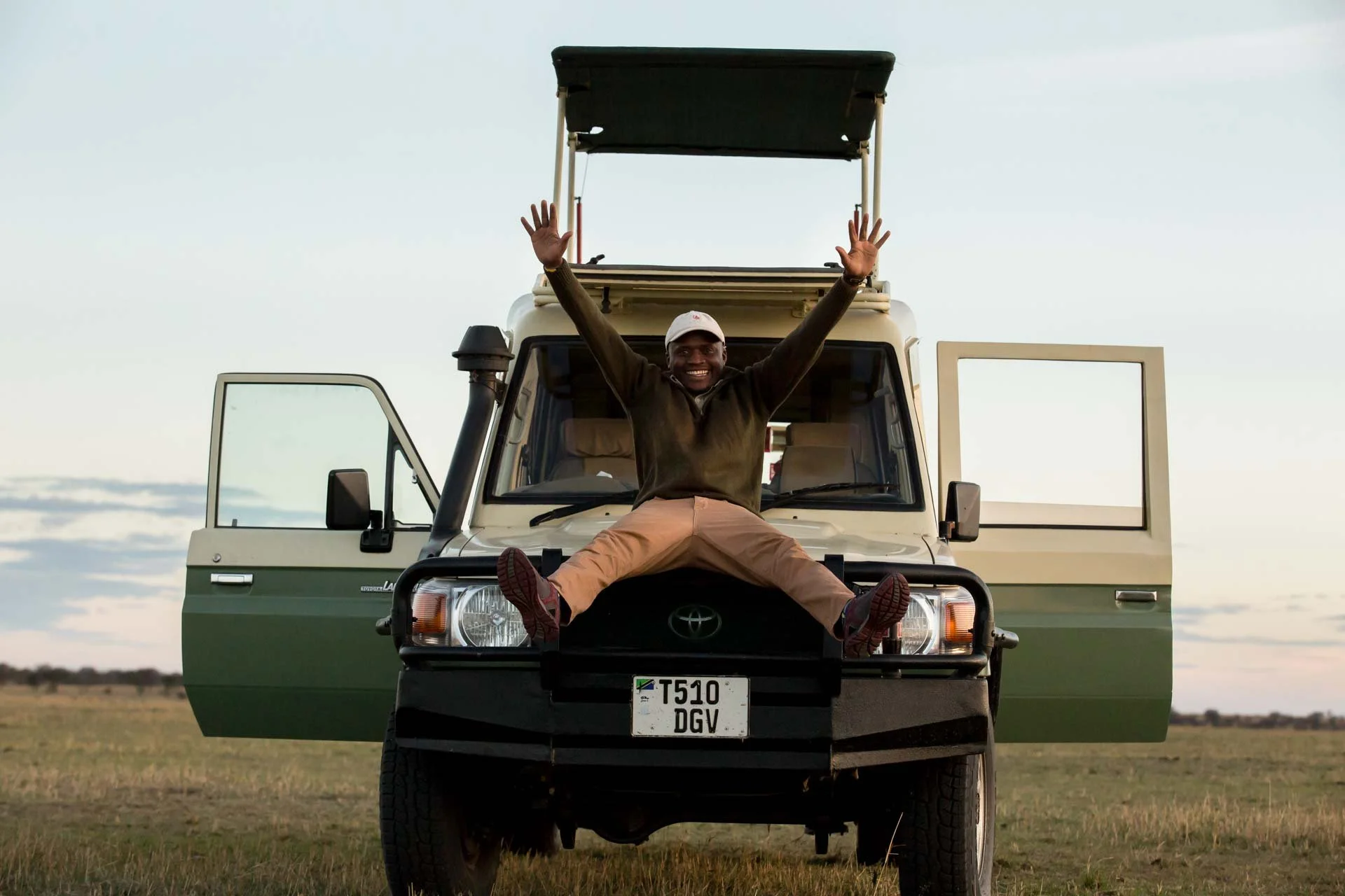 A man sitting on the front of a Toyota Land Cruiser, smiling and with arms raised, with open doors and a roof hatch open in an open grassy field during sunset or sunrise.