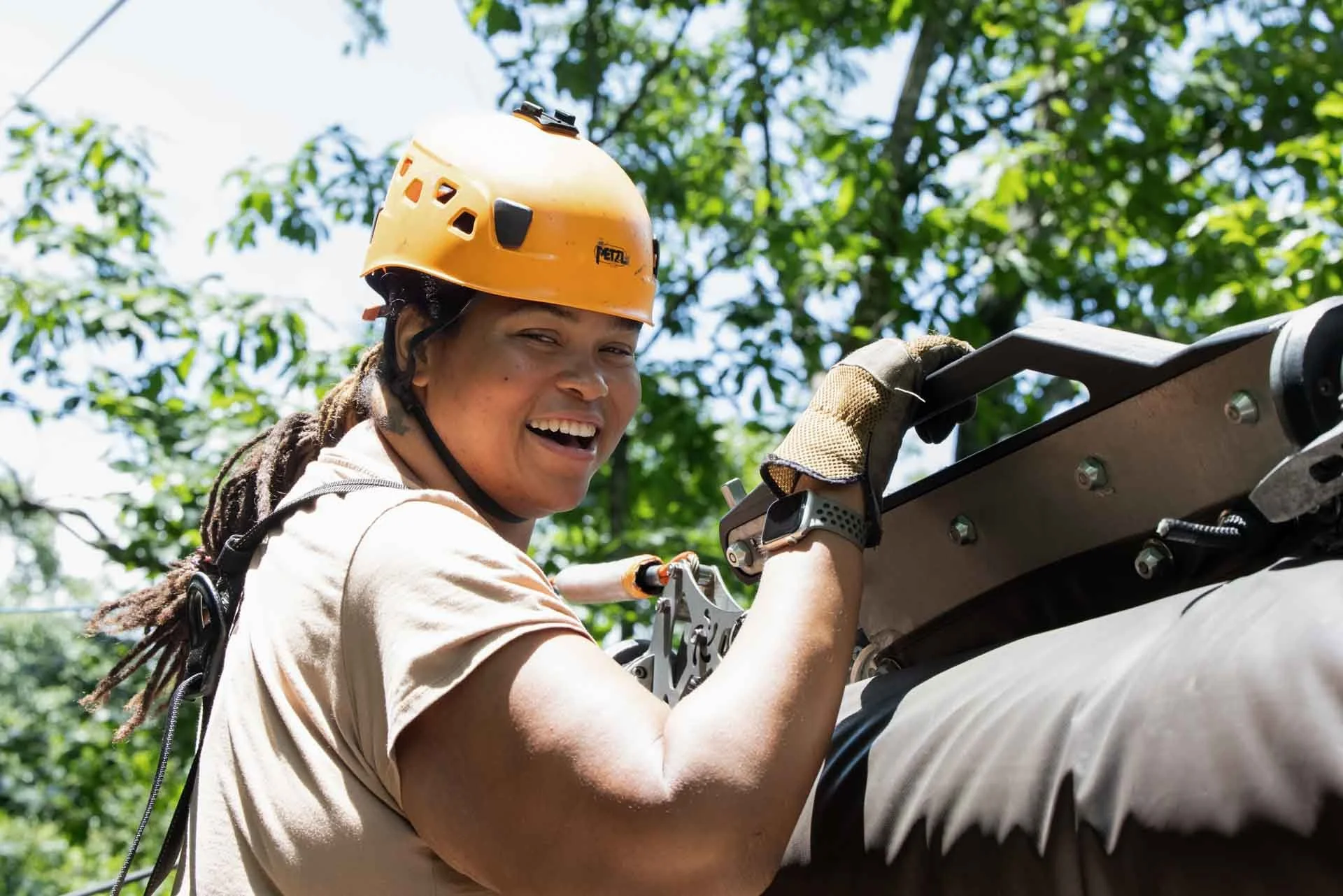 A woman wearing an orange helmet, gloves, and beige clothing working on climbing equipment outdoors with green trees in the background.