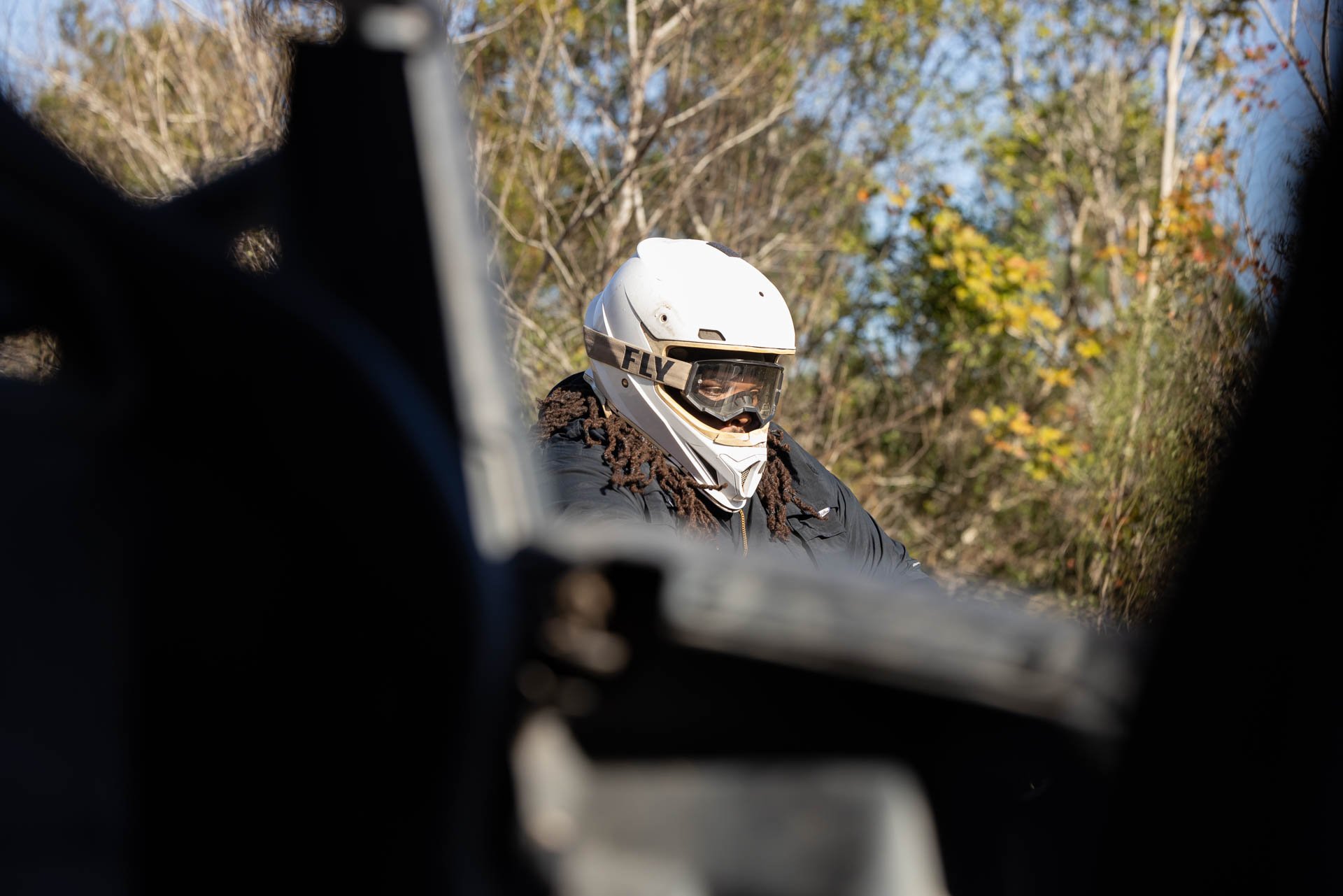 Person wearing a white motorcycle helmet and goggles, dressed in a black jacket, seen through a motorcycle's frame, with a background of trees and blue sky.