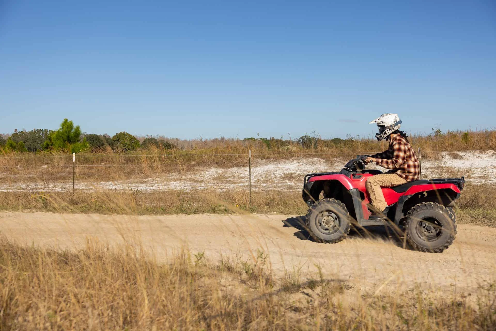 Person riding a red ATV on a sandy trail in an open field with grass and sparse bushes, wearing a helmet and a plaid shirt.