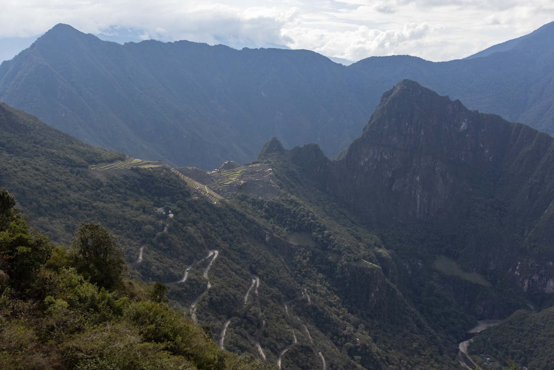 Mountain landscape with winding roads, lush green hills, and misty mountain peaks in the background under cloudy sky.