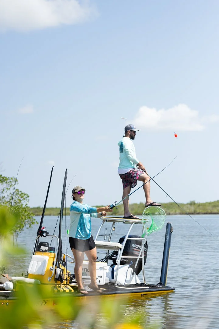 Two people fishing on a small boat in a calm body of water under a partly cloudy sky. One person is standing on a raised platform, reeling in a fish, while the other person is holding a fishing rod. The boat has fishing gear and equipment on board.