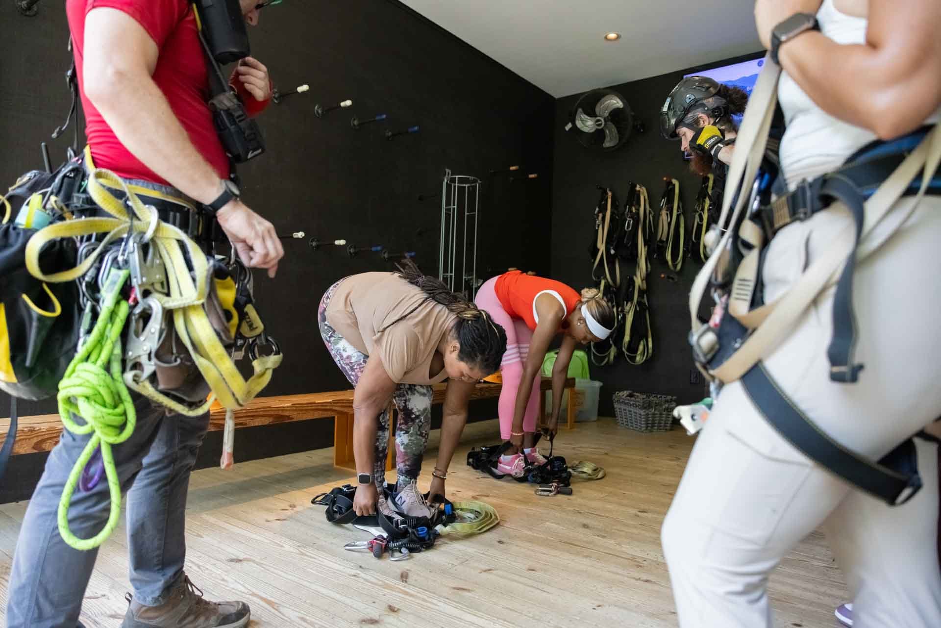 Two women are bending over on a wooden floor to put on climbing harnesses, while two men stand nearby observing. The room has climbing gear and safety equipment hanging on the dark wall and placed on the floor.