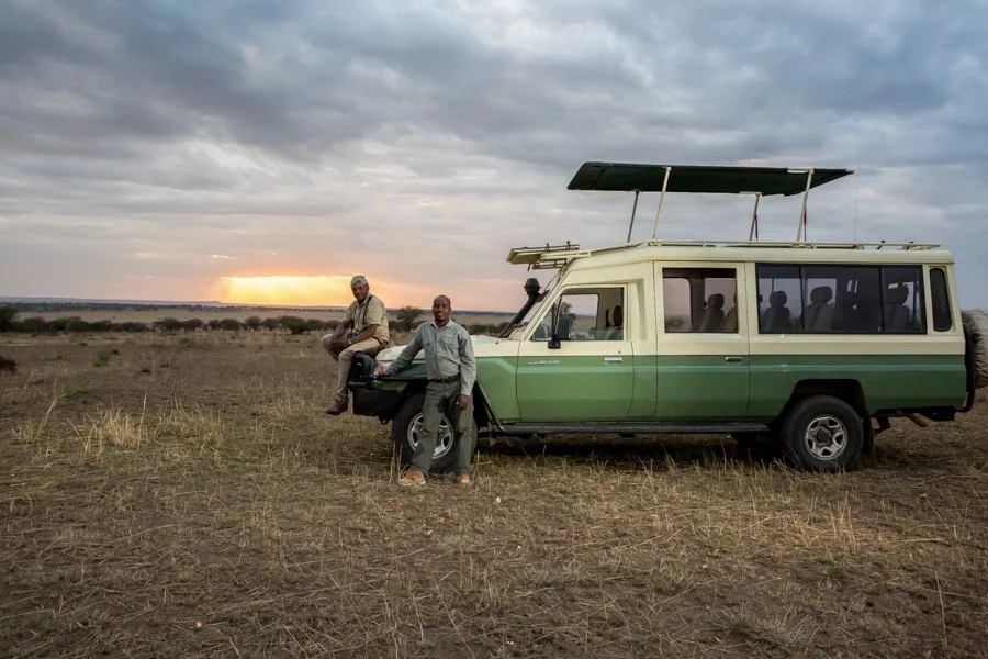 Two men with a safari vehicle in an open grassy landscape during sunset, one sitting on the vehicle's front and the other standing beside it.