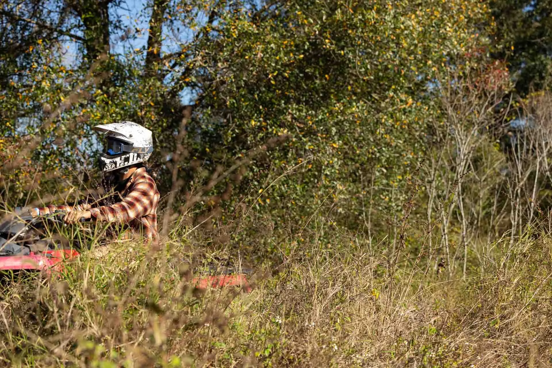 A person riding a quad bike through tall grass and bushes in a natural outdoor setting, wearing a helmet and a plaid shirt.