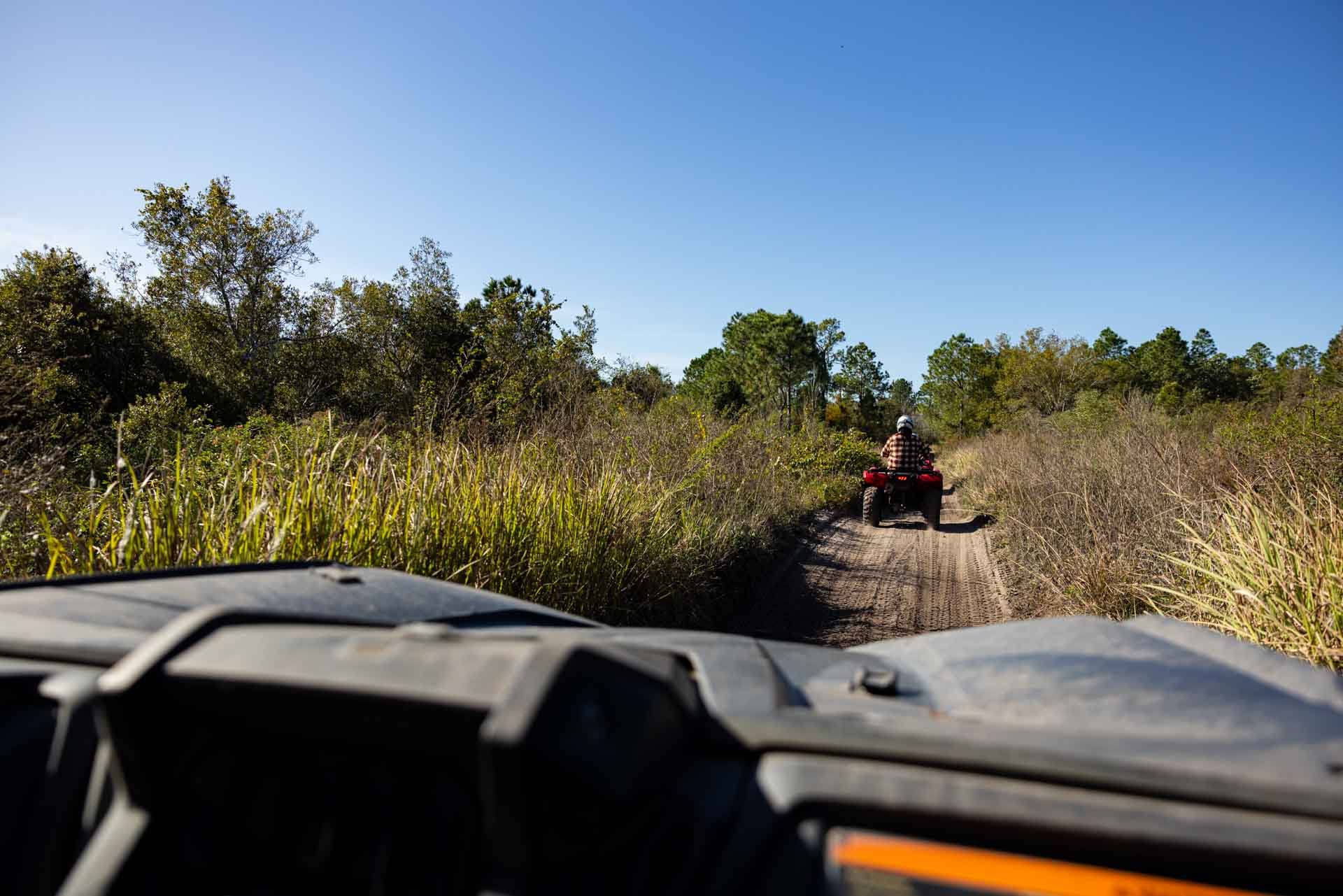 View from inside a vehicle on a dirt trail with tall grass on either side, leading to an ATV ahead with a person wearing a helmet riding on the trail under a clear blue sky.