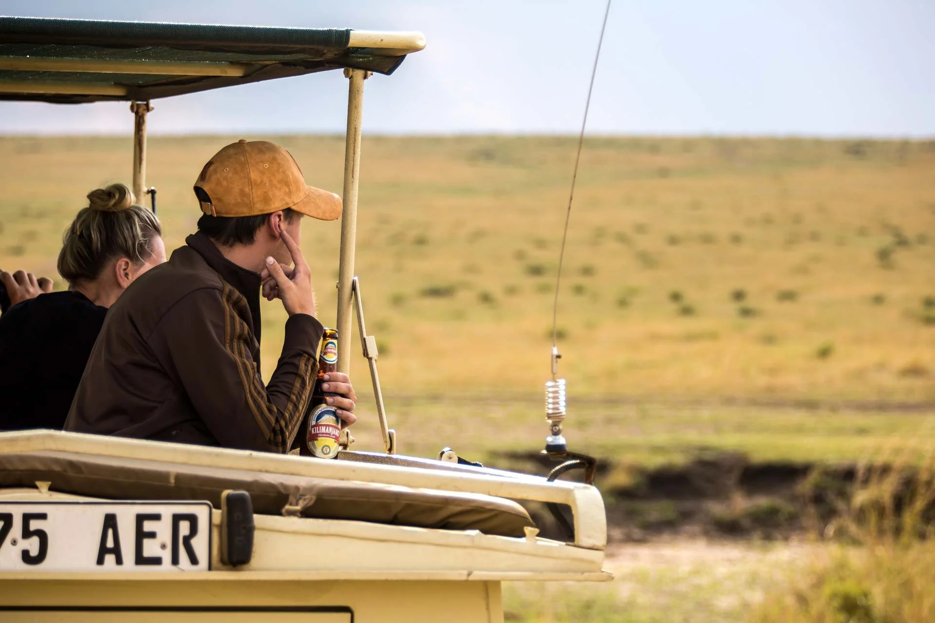 Two people are sitting in the back of a safari vehicle in an open field, observing the landscape. One person wears a brown jacket and a tan cap, holding a bottled beverage, and appears to be deep in thought. The other person has blonde hair tied up a