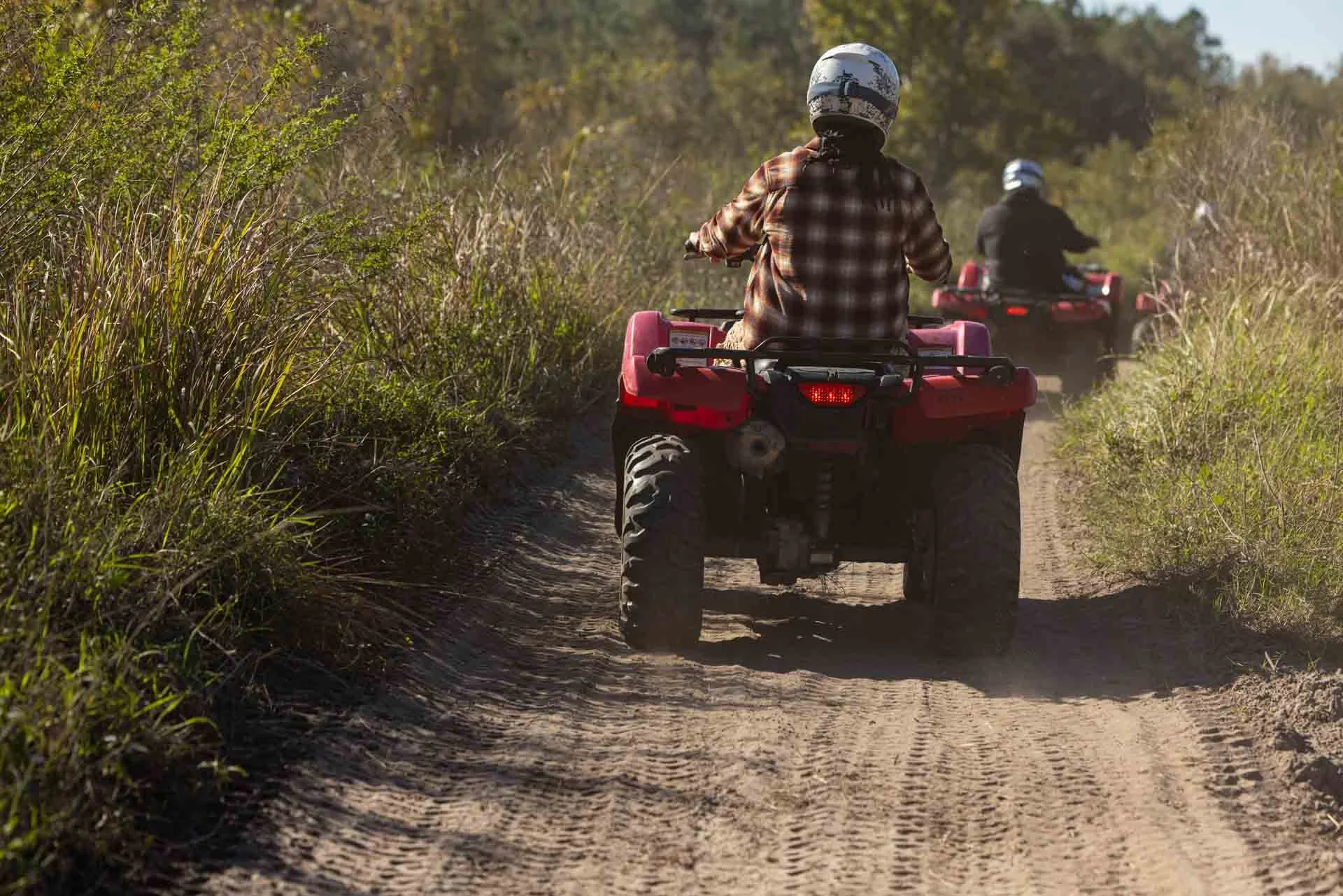 Two people riding all-terrain vehicles (ATVs) on a dirt trail through tall grass and trees during daytime.