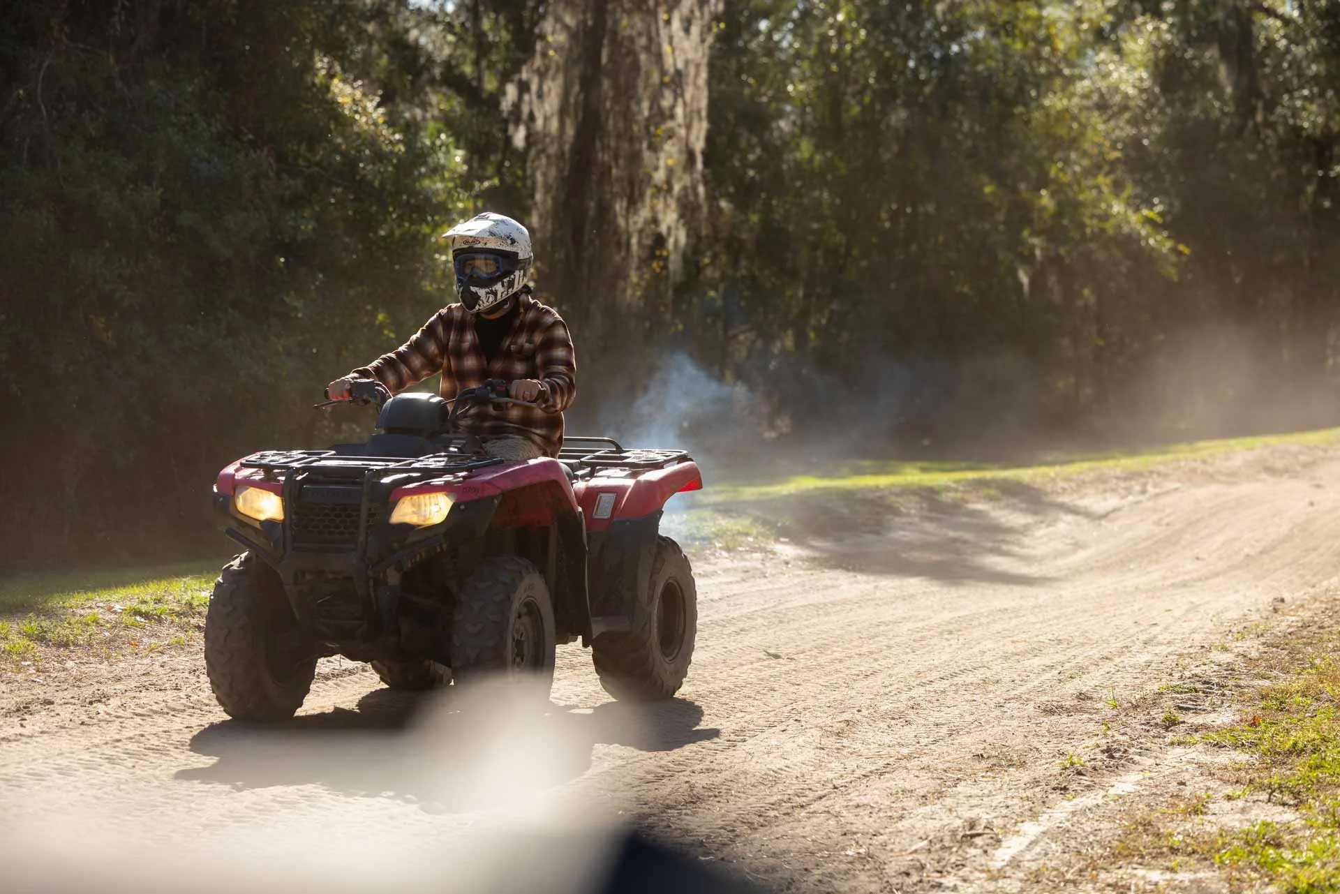 A person riding an ATV on a dirt trail through a wooded area, wearing a helmet and a plaid shirt.