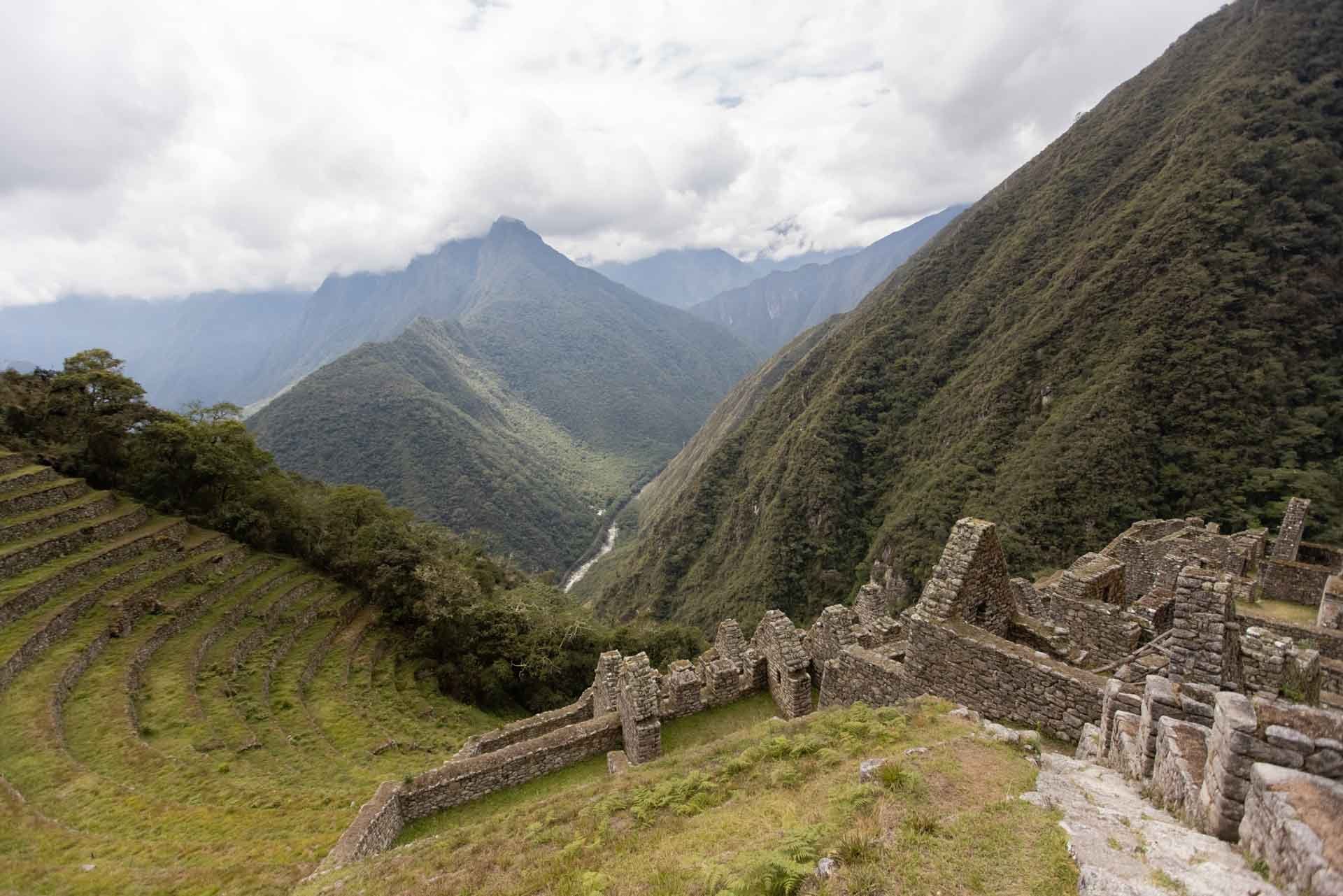 Ancient Incan ruins with stone walls and terraces in a lush mountain valley, overlooking steep green mountains and a cloudy sky.