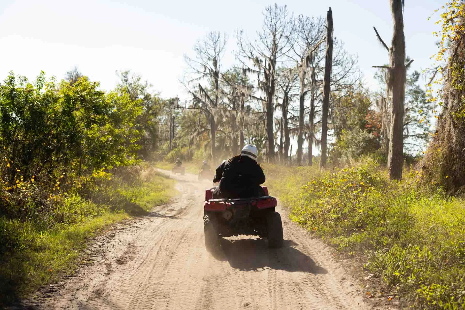 Person riding a quad bike on a dirt trail through a wooded area with tall trees and green bushes.