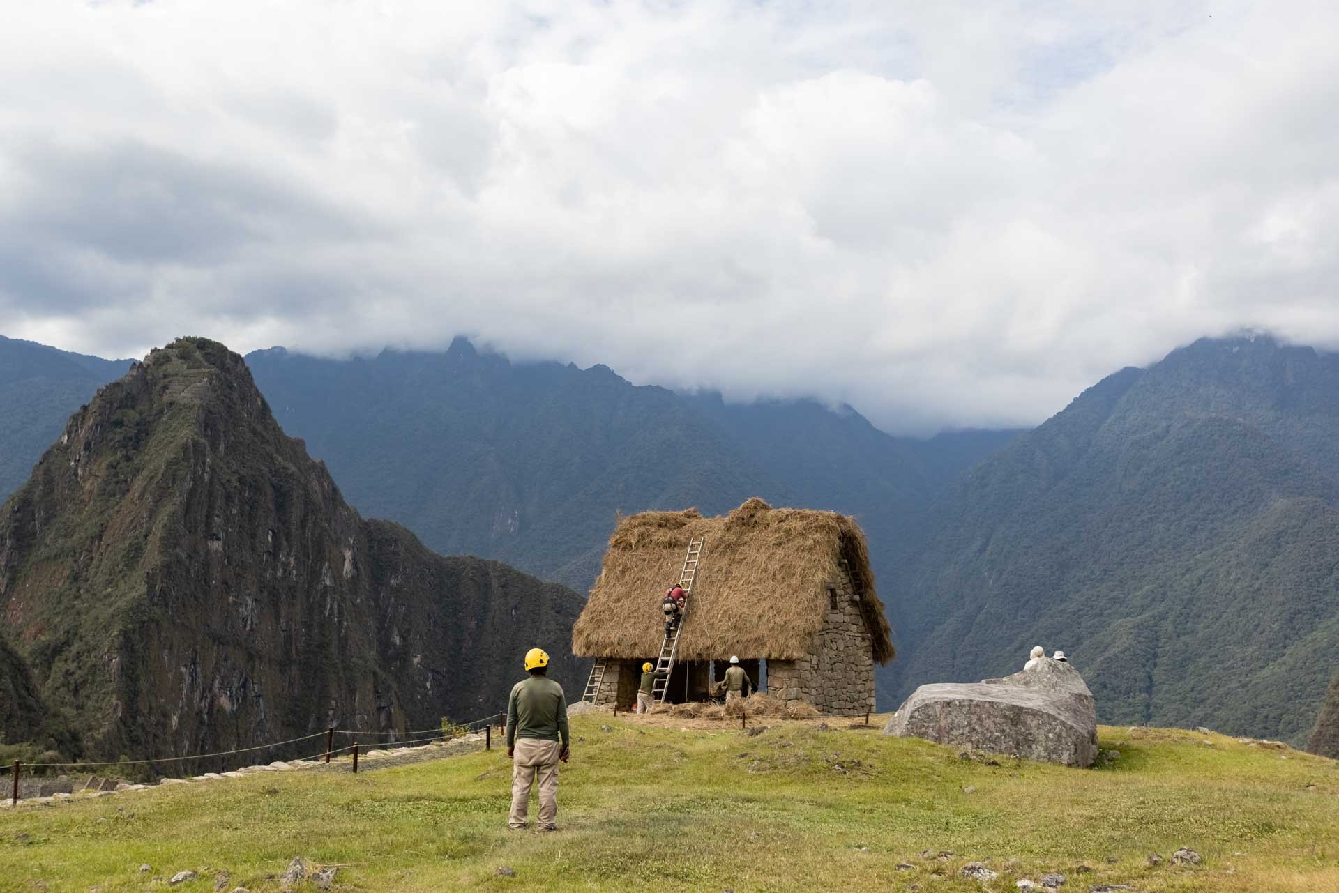Researchers working on a stone and thatched roof building at Machu Picchu, Peru, with mountains and cloudy sky in background.