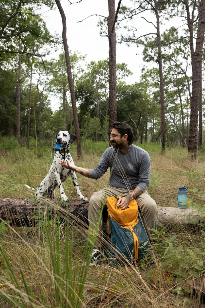 A man with dark hair and a beard, wearing glasses, a gray long sleeve shirt, and khaki pants, sits on a log in a forested area with tall trees and grass. He is smiling and gently touching the paw of a Dalmatian dog holding a camera in its mouth. Ther
