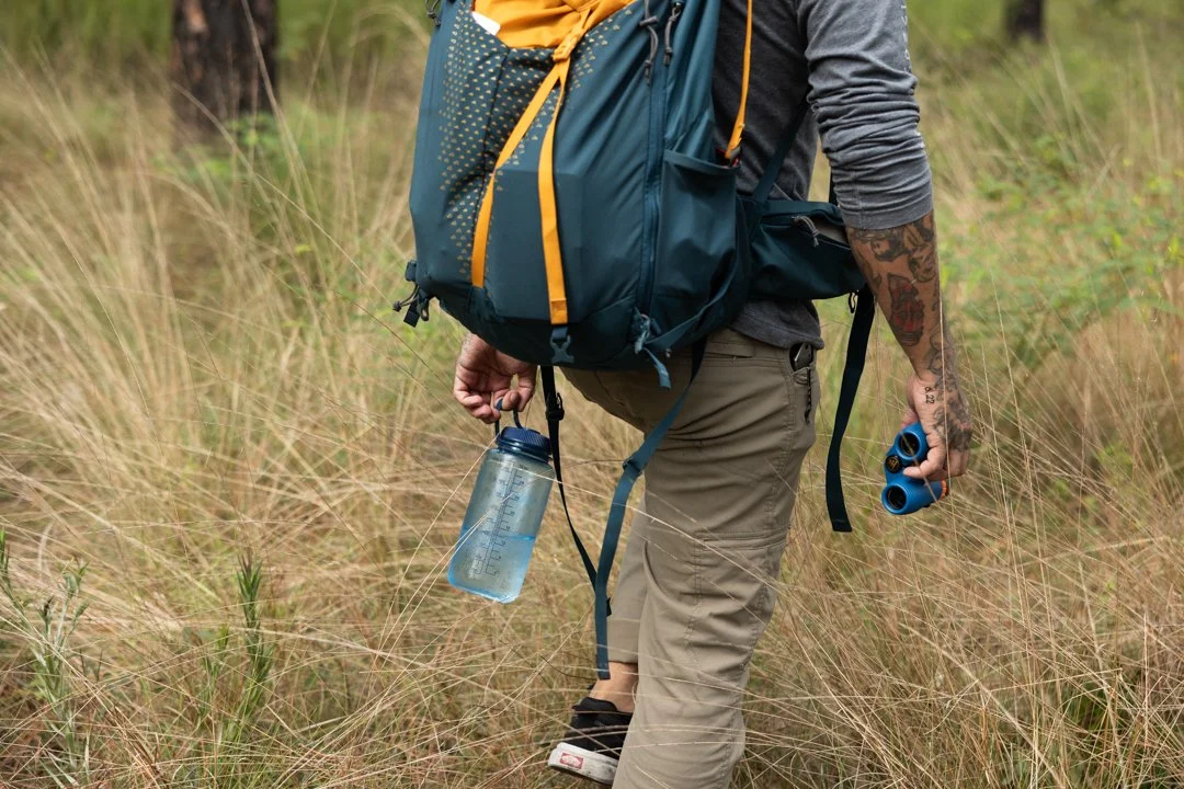 A person wearing khaki pants and a gray long-sleeve shirt hiking through tall grass carrying a blue backpack, water bottle, and blue binoculars.