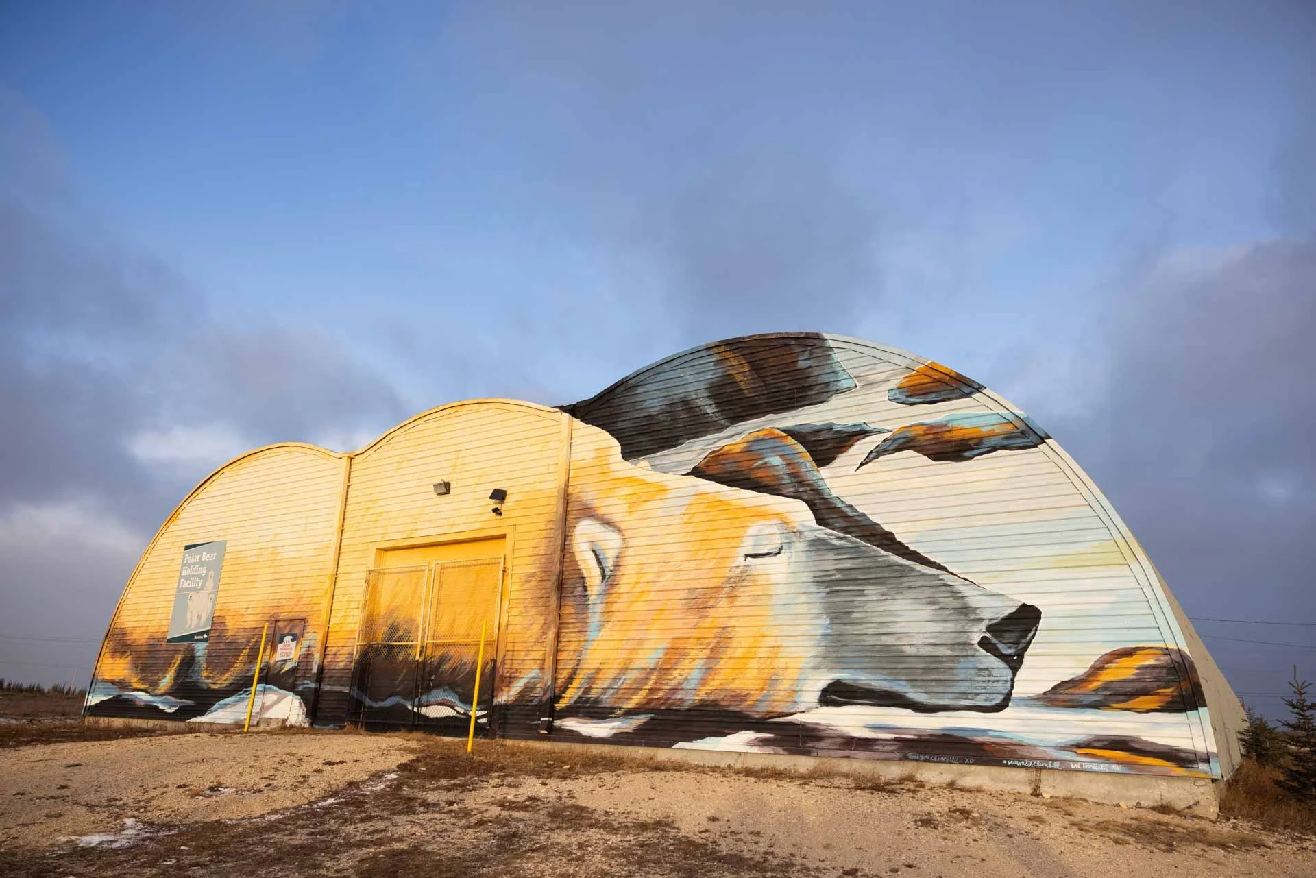 A large mural of a sleeping dog painted on a Quonset hut, with a landscape background and clouds in the sky.