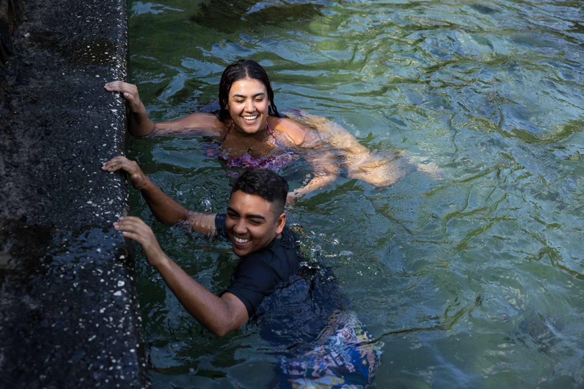 Two smiling people holding onto a stone ledge while partially submerged in a body of water. One woman and one man are visible, with the woman in a purple swimsuit and the man in a dark shirt. The water is greenish with ripples reflecting sunlight.