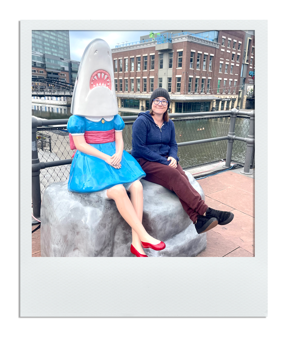 A woman sitting next to a cartoonish shark statue with a human body, on a stone bench near a river in an urban area.
