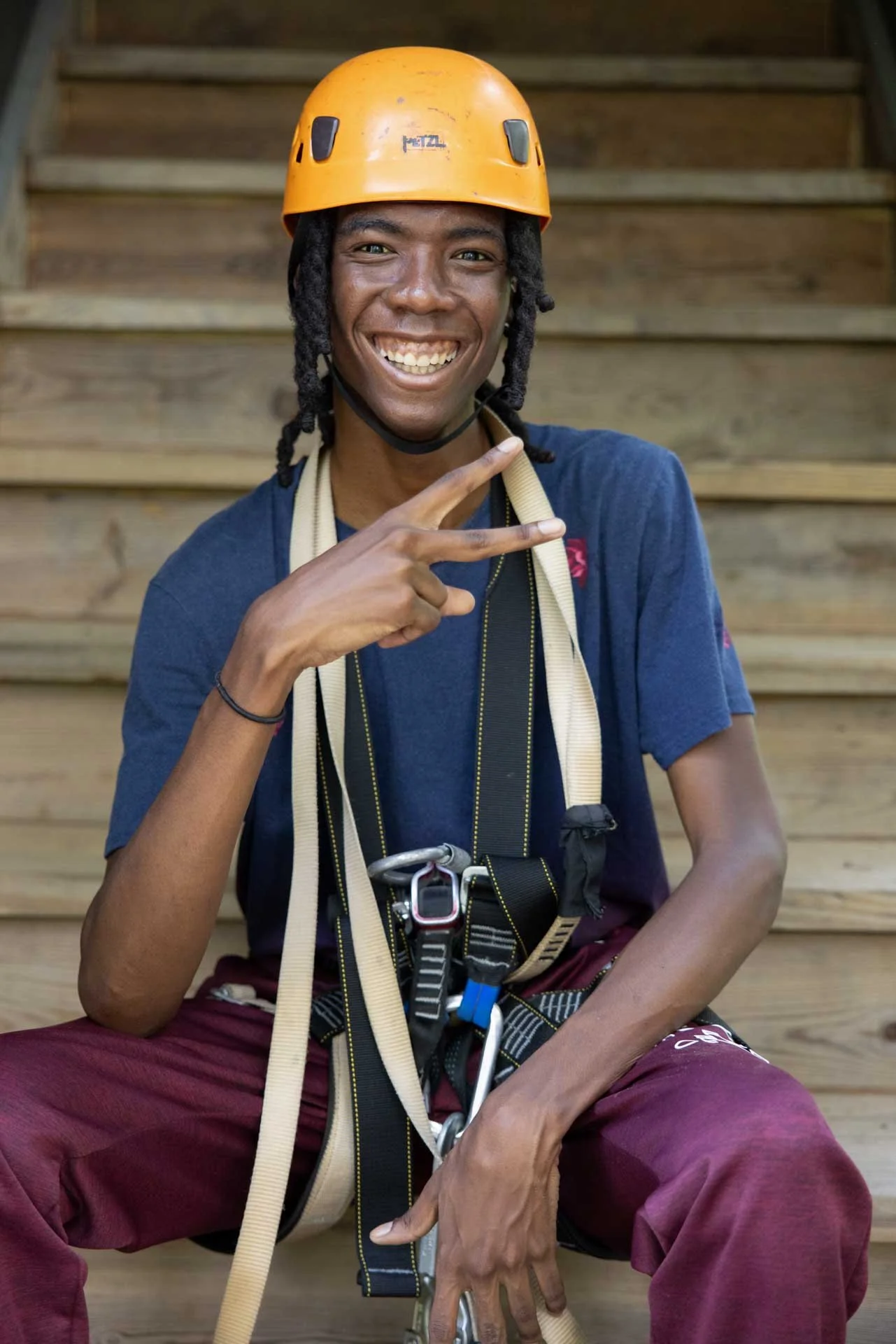 A young man smiling, wearing an orange climbing helmet, sitting on wooden stairs, with climbing harness and gear.
