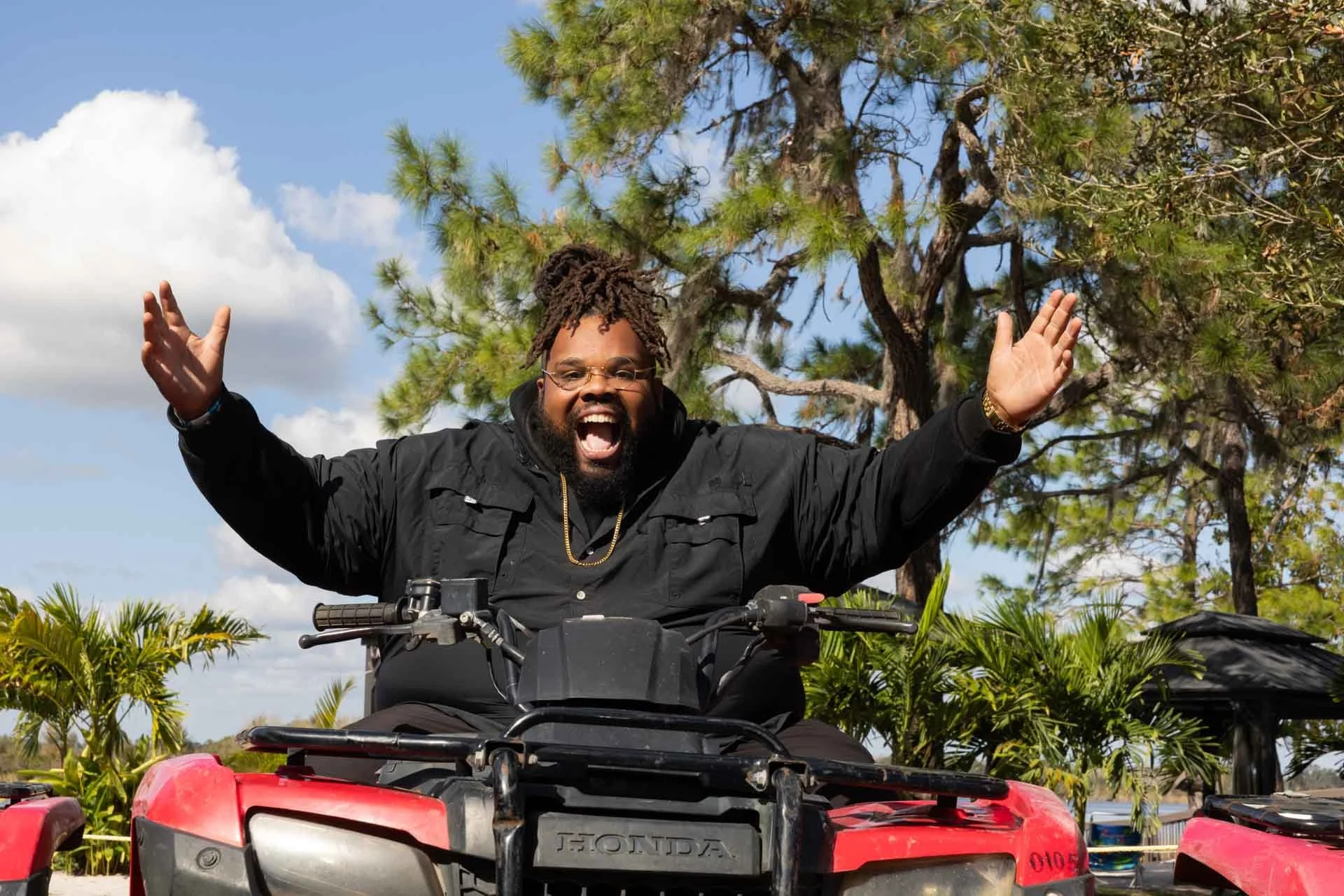 A man with glasses and a beard, wearing a black jacket, rides a red all-terrain vehicle (ATV) with his arms raised in celebration, outdoors with trees and blue sky in the background.