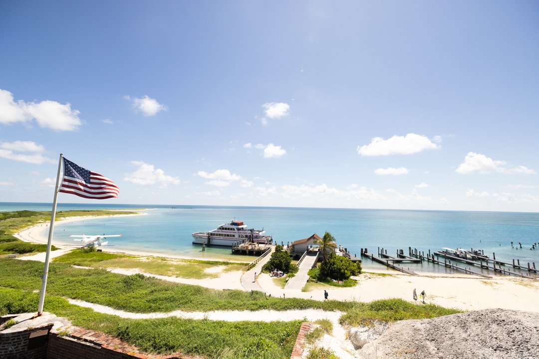 A scenic view of a harbor with boats and a dock, an American flag on a flagpole, sandy beach, clear blue water, and a partly cloudy sky.