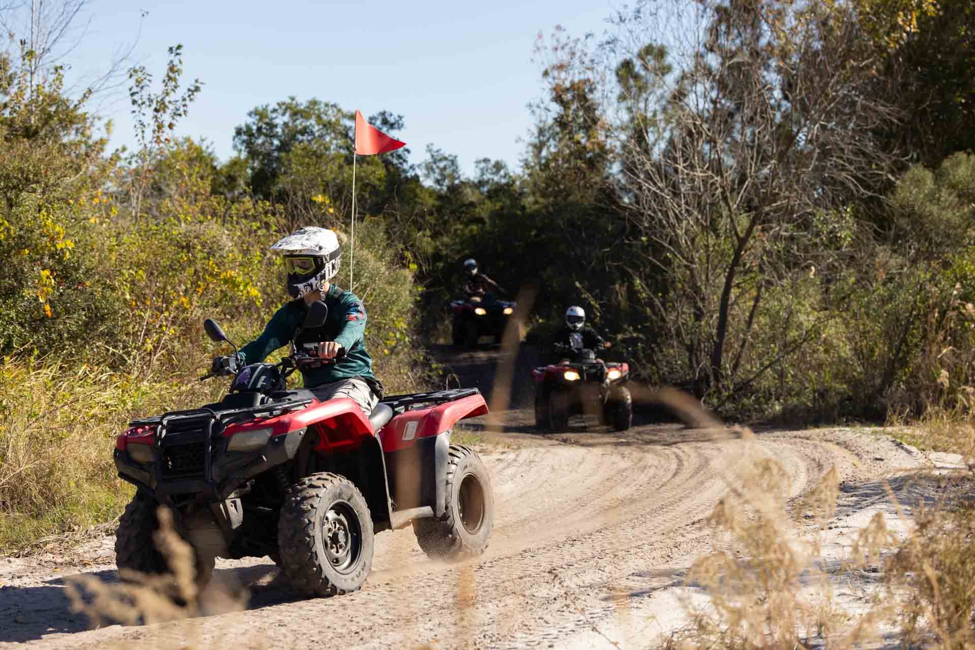Three people riding all-terrain vehicles (ATVs) on a dirt trail surrounded by trees and bushes during daytime.