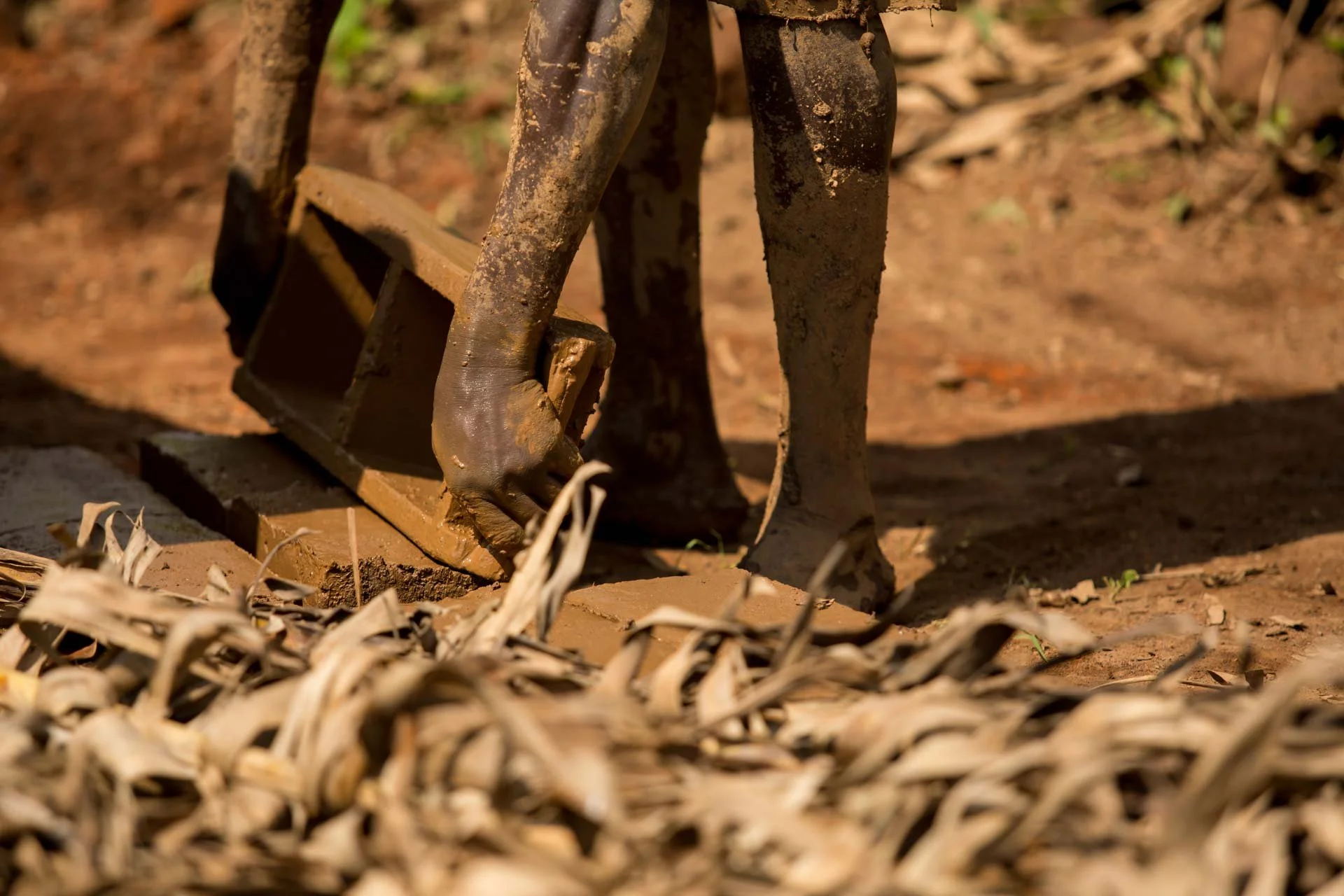 Close-up of a person's muddy hands using a blunt axe to chop wood outdoors, with dry leaves and soil on the ground.