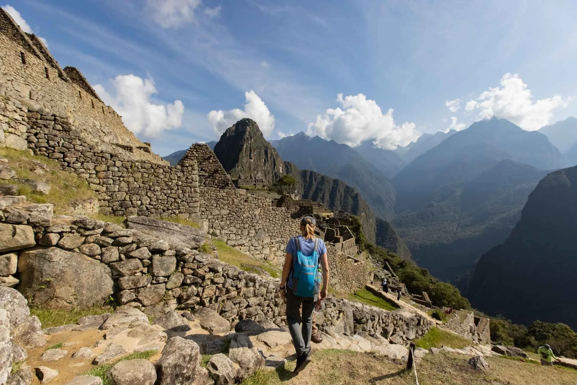 A woman with a blue backpack and cap walking on stone steps in front of ancient Incan ruins, Machu Picchu, with mountains and clouds in the background.