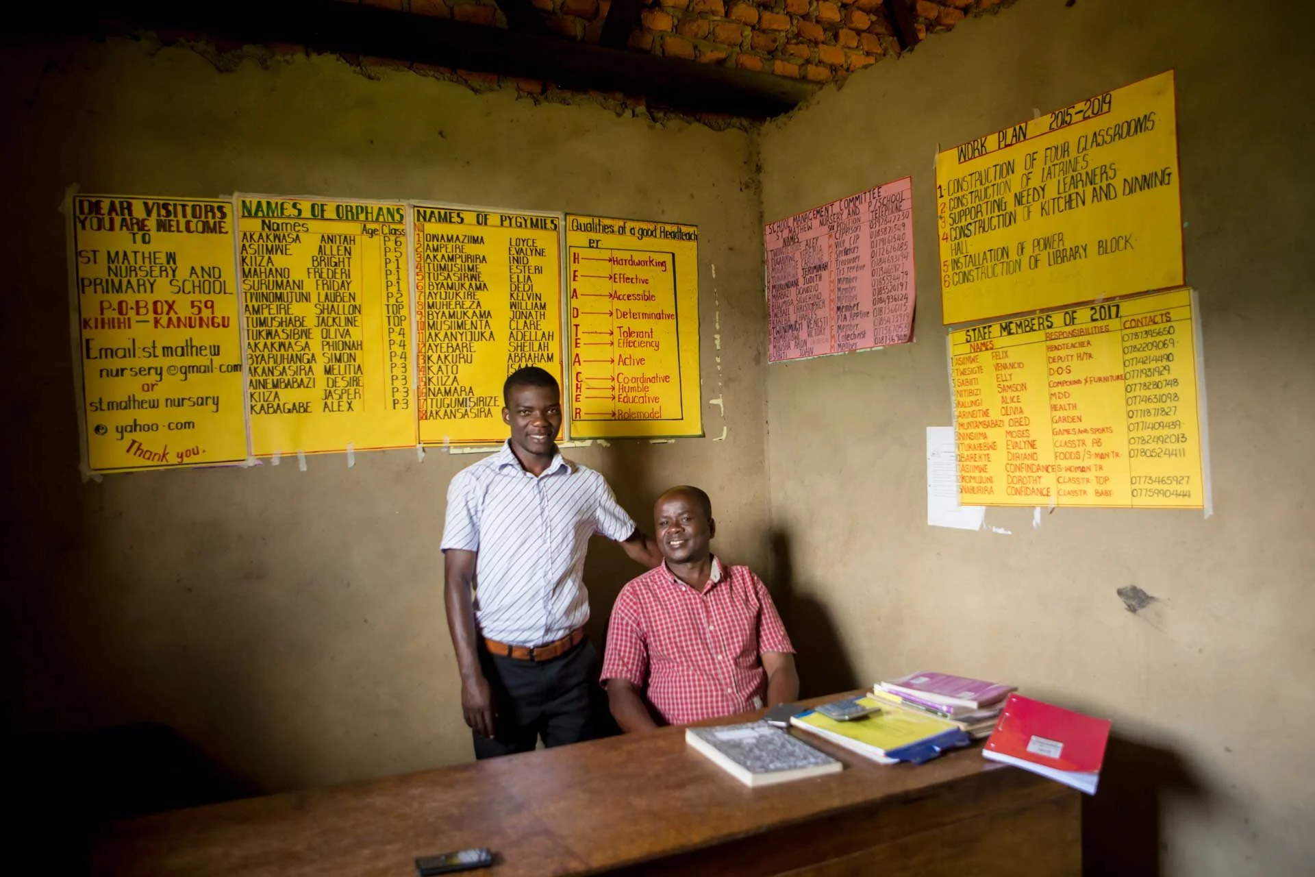 Two men in a classroom with educational posters on the wall. One man is standing, and the other is sitting at a desk with books and notebooks.