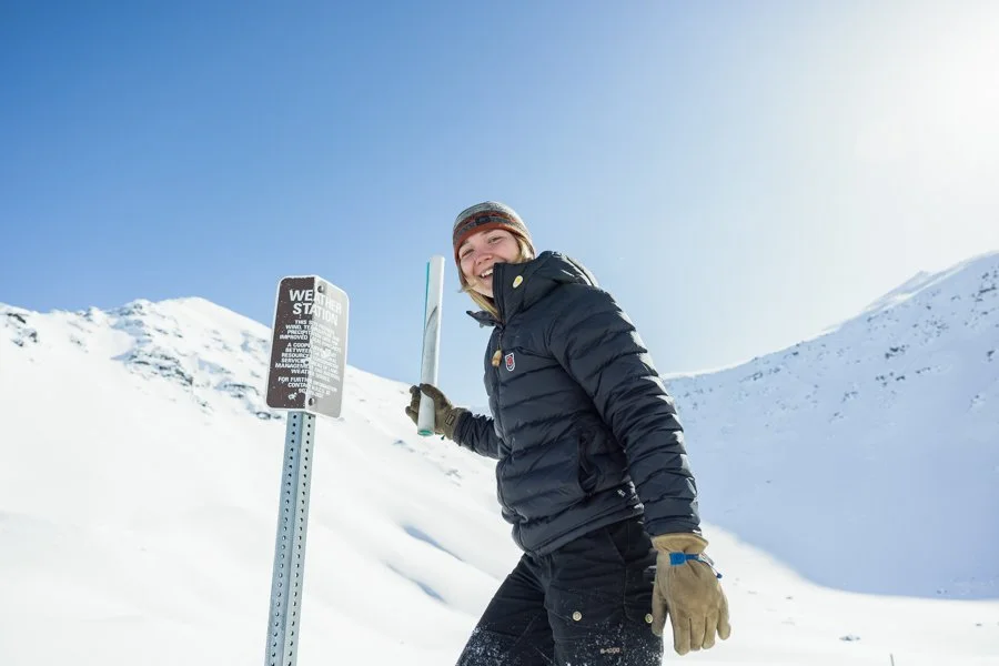 A smiling woman in winter gear standing in a snowy mountain landscape, holding a snow shovel, with a weather warning sign nearby.