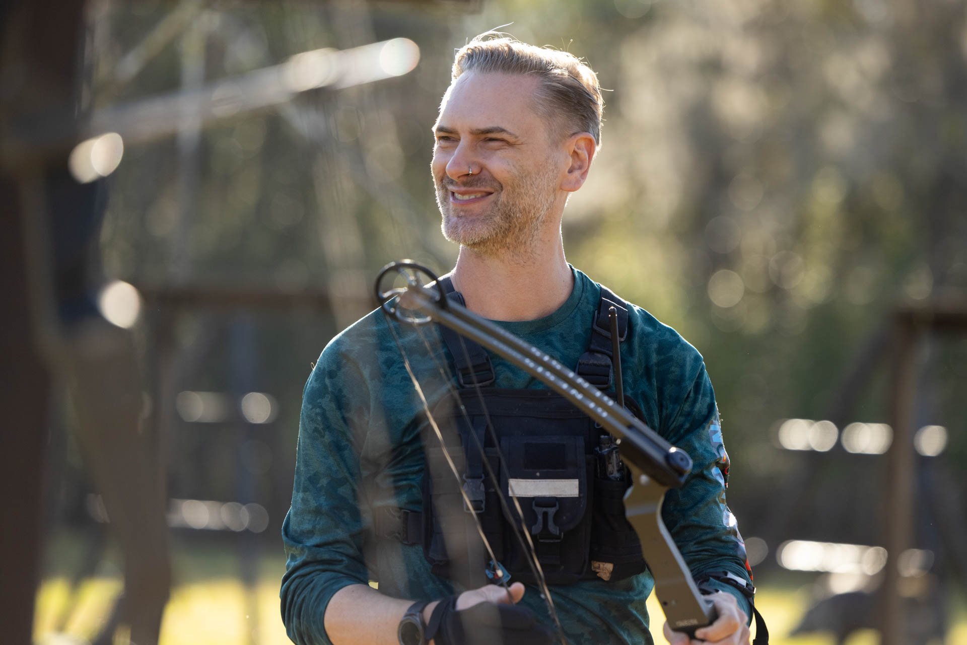 A man with gray hair and a beard smiling outdoors, holding a metal detector, wearing a dark vest and gloves, with trees and sunlight in the background.