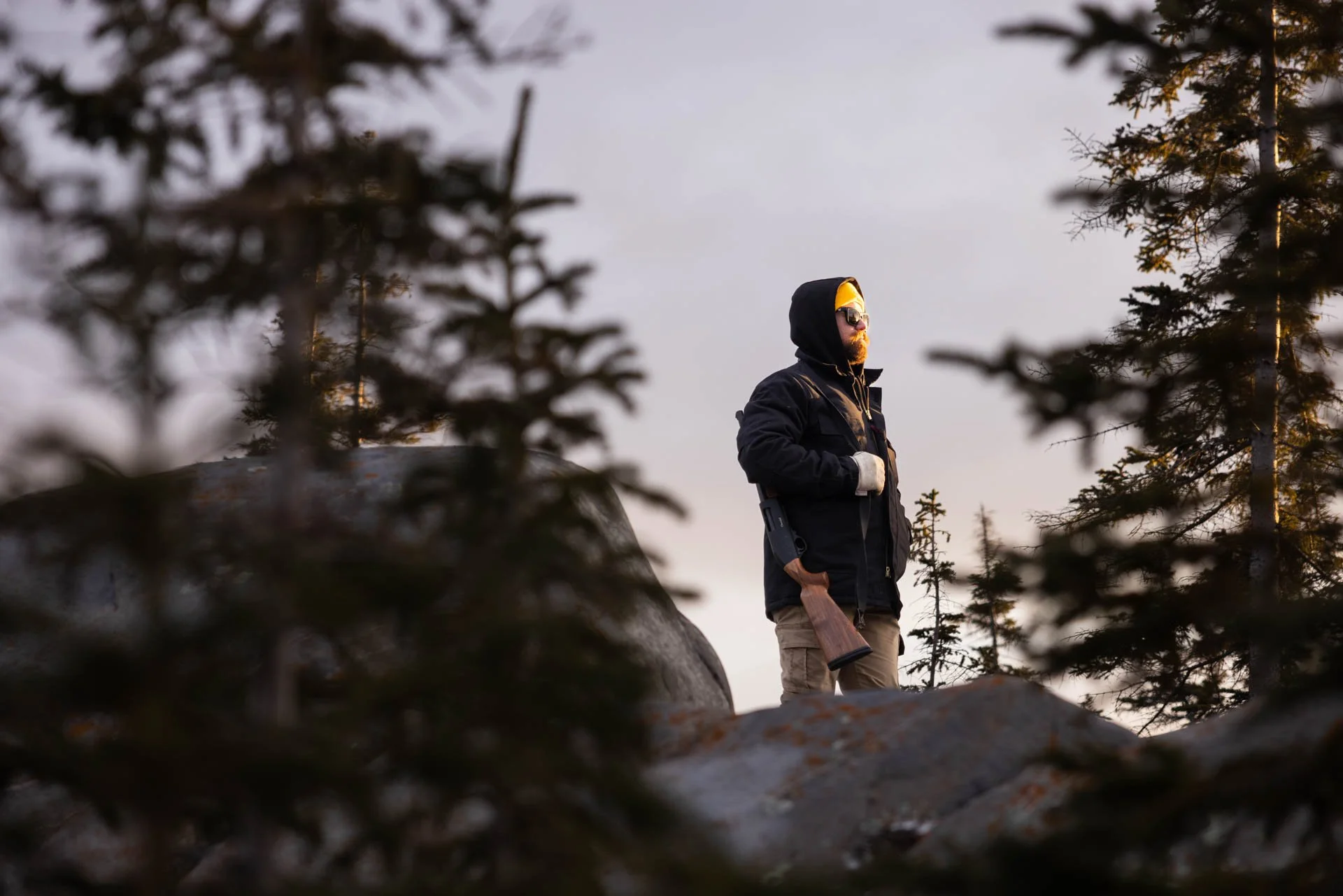 A man standing on rocks surrounded by trees, wearing sunglasses, a hoodie, gloves, and a jacket, holding a rifle.