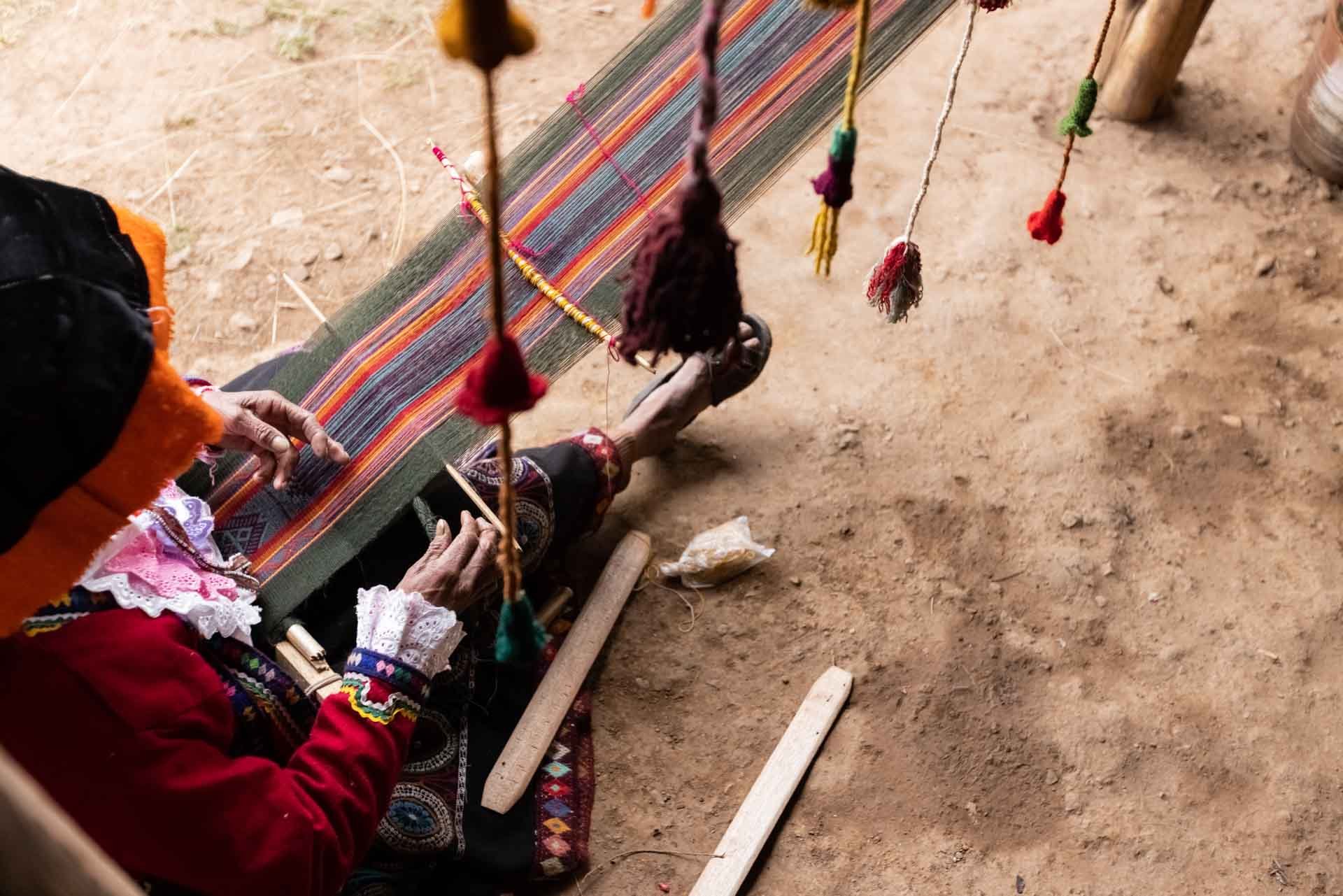 A person weaving traditional colorful textile on a loom outdoors, with hanging decorative tassels.