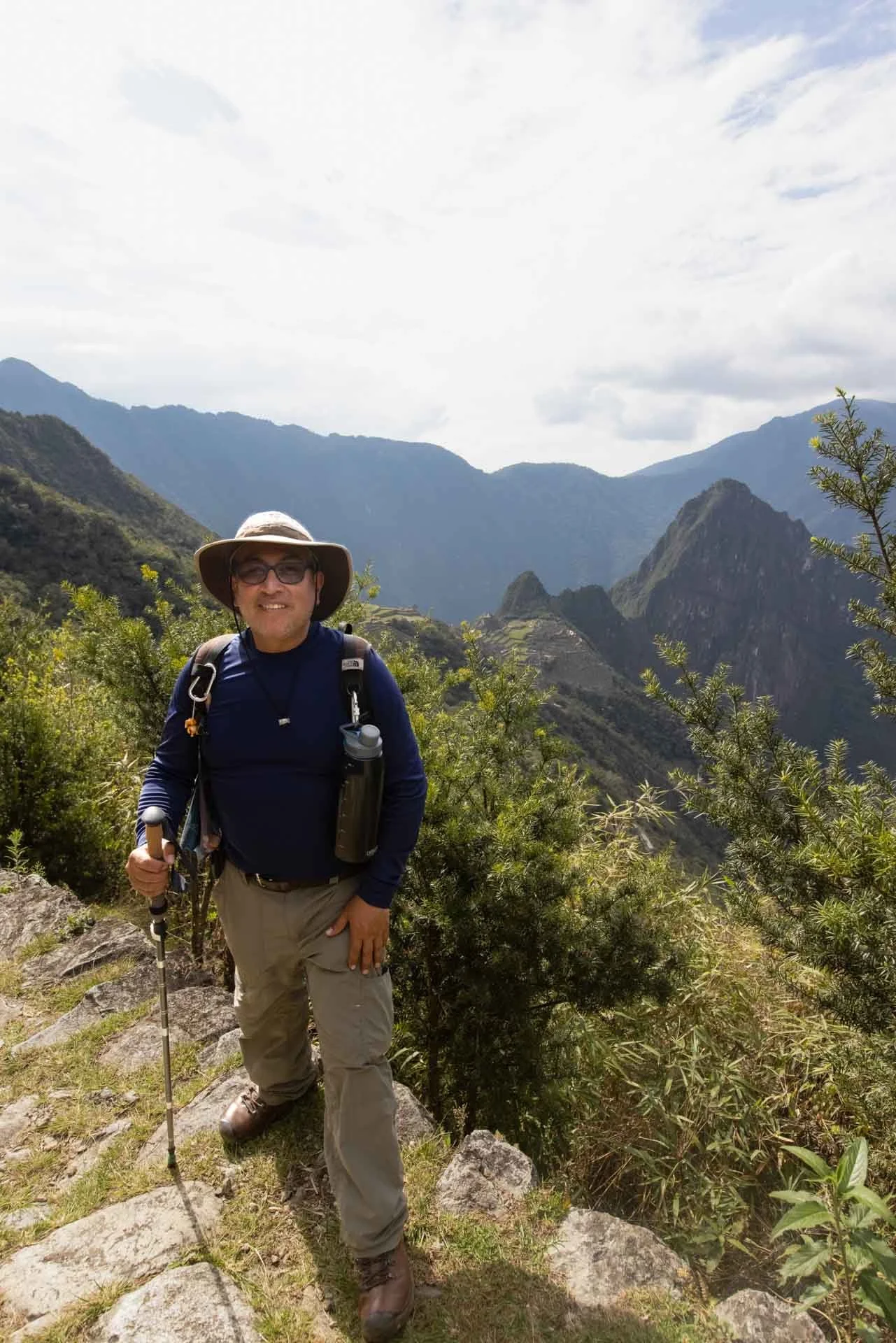 A person dressed for hiking in front of mountainous landscape with lush green vegetation and cloudy sky.