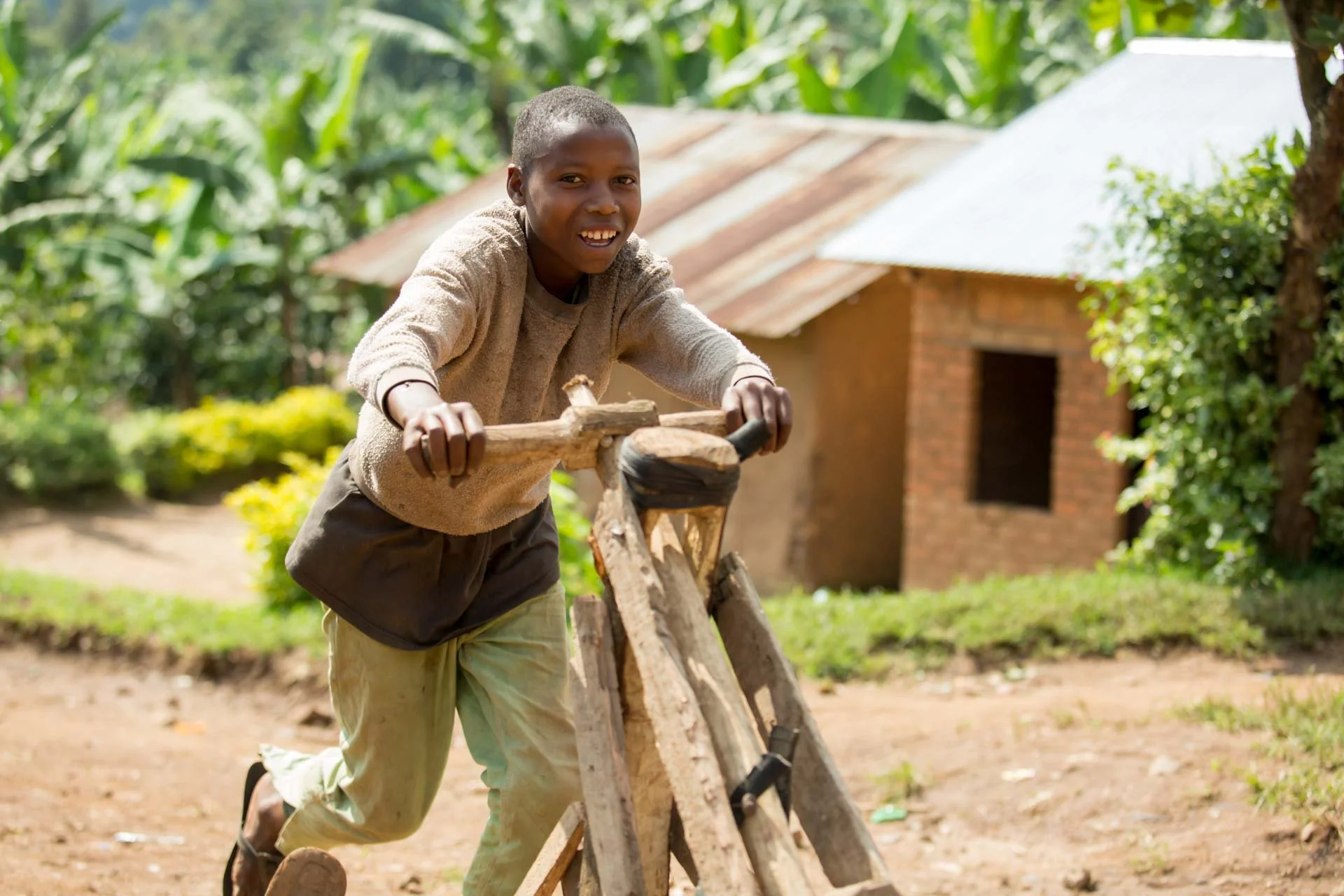 A young boy outdoors, riding a wooden scooter with a rustic building and green foliage in the background.