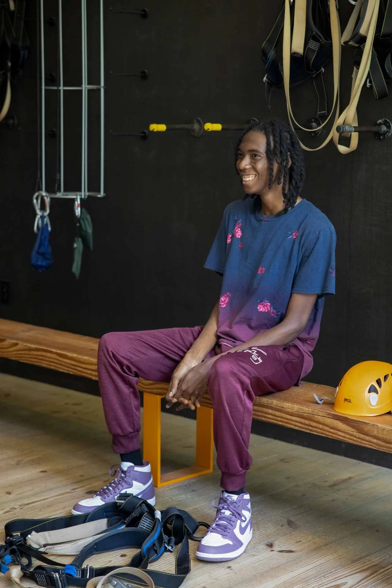 A young man wearing purple and white sneakers, maroon pants, and a blue shirt with pink flowers, sitting on a wooden bench in a rock climbing gym, smiling. Climbing gear including a helmet and harness is nearby, with rock climbing holds, ropes, and e