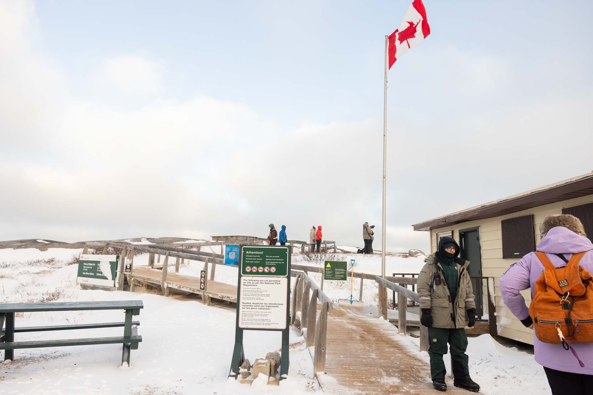 People visiting a snowy area near a wooden lookout platform with a Canadian flag flying high, and signs with rules posted in front, during a cloudy day.