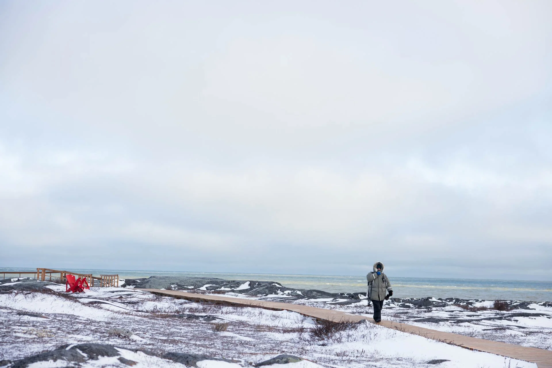 A person walking on a wooden pathway along a rocky, snow-covered coastline under a cloudy sky, with a red chair and a small fence nearby.