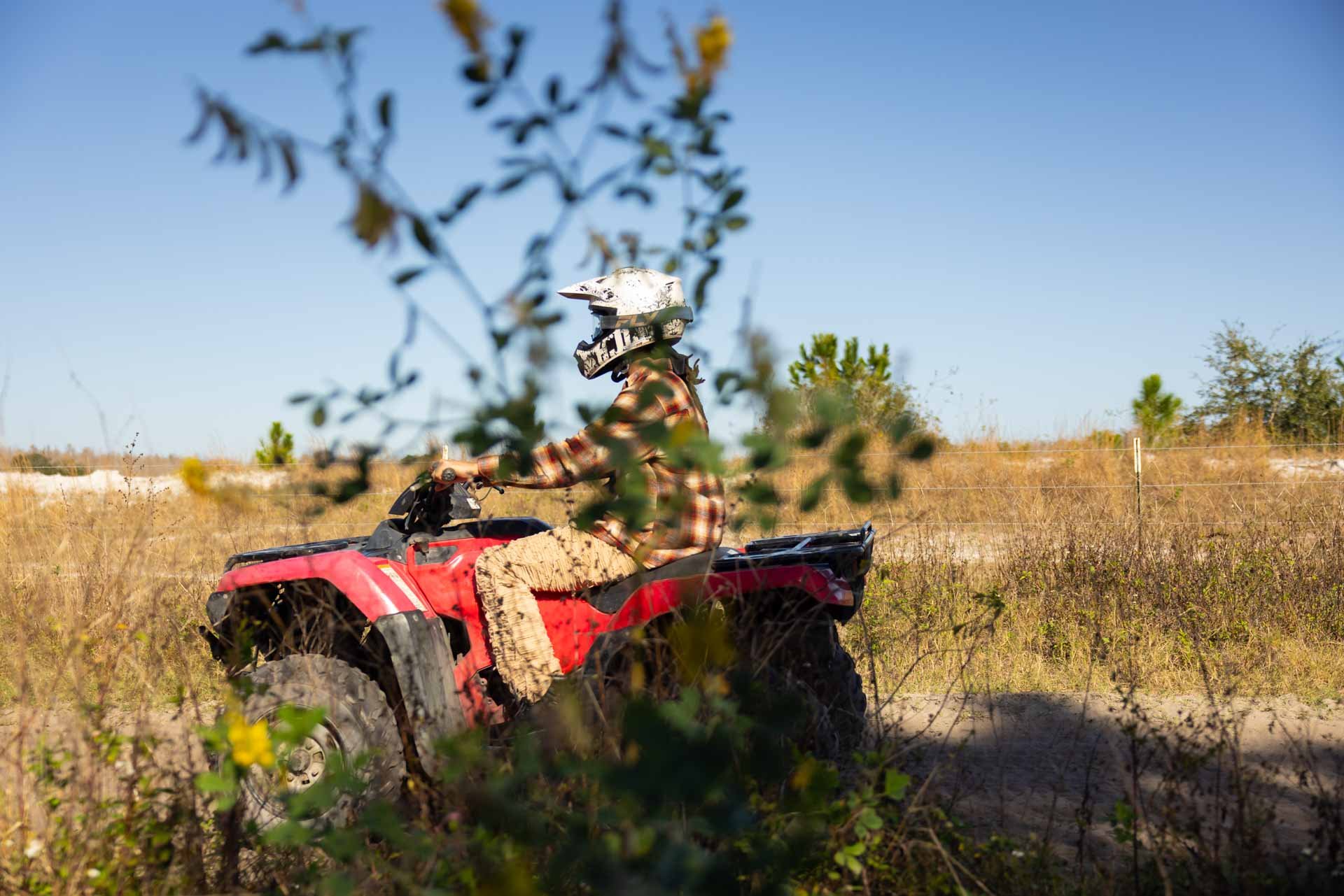 Person in a checkered shirt and camouflage pants riding a red all-terrain vehicle (ATV) wearing a white helmet in a dry, open field with sparse trees and bushes, viewed through some foliage.