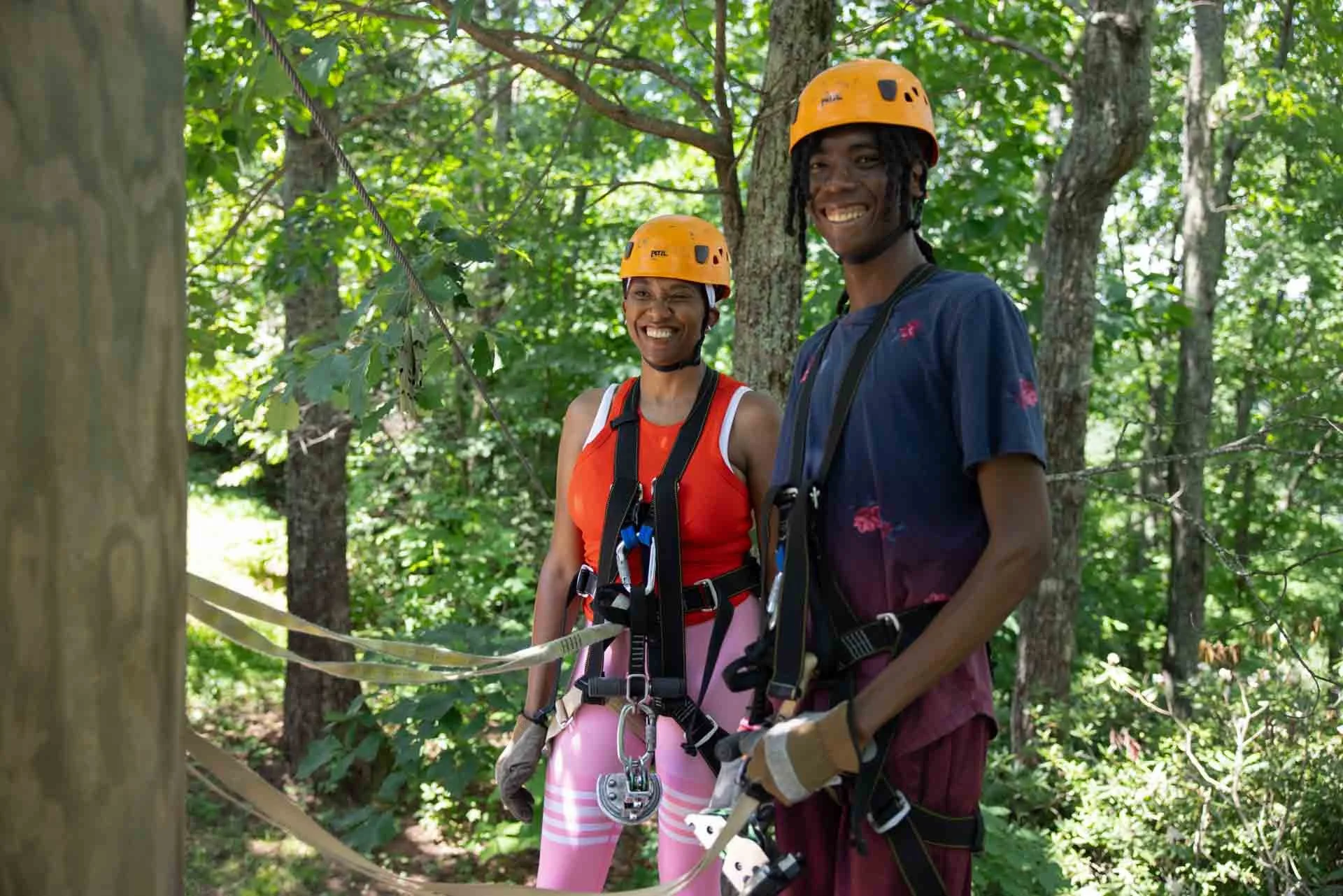 Two smiling people wearing safety harnesses and helmets in a forest, preparing for zip lining or tree climbing activity.