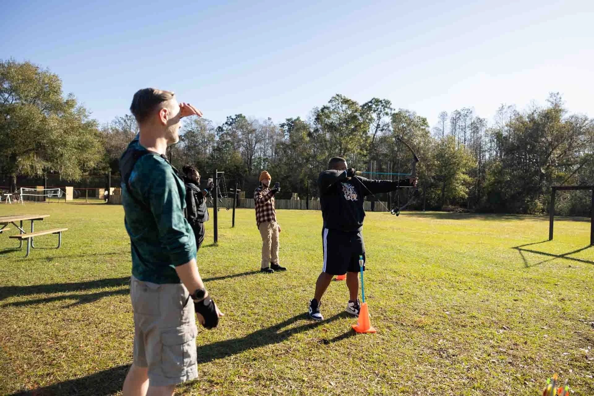 A group of five young men outdoors on a bright, sunny day, with one man practicing archery aiming at a target, while the others watch and take photos.