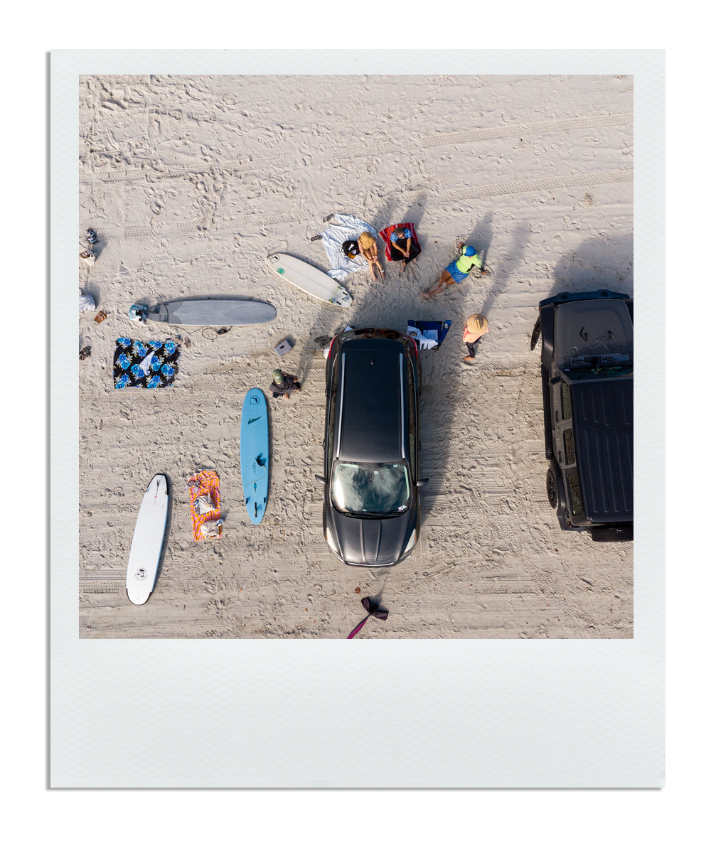 An aerial view of a beach scene with surfboards, a blanket, and people gathered near parked cars on sandy beach.