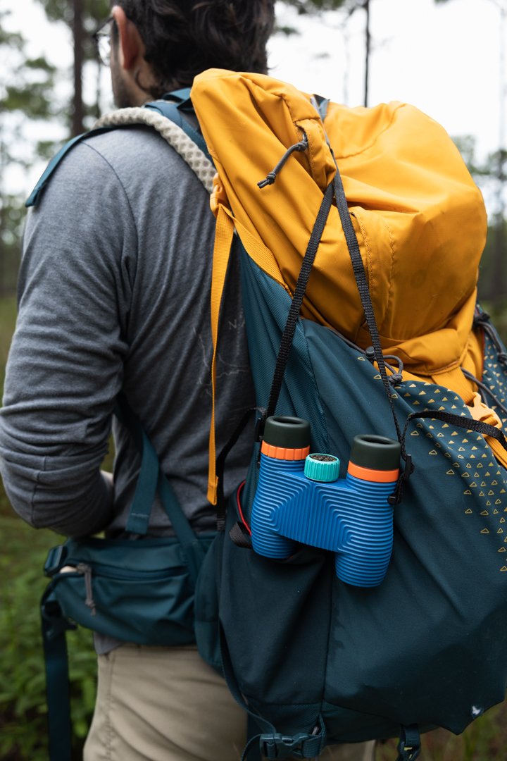 A person wearing a gray shirt and khaki pants with a yellow and blue hiking backpack, holding binoculars attached to the side of the backpack, outdoors in a forested area.
